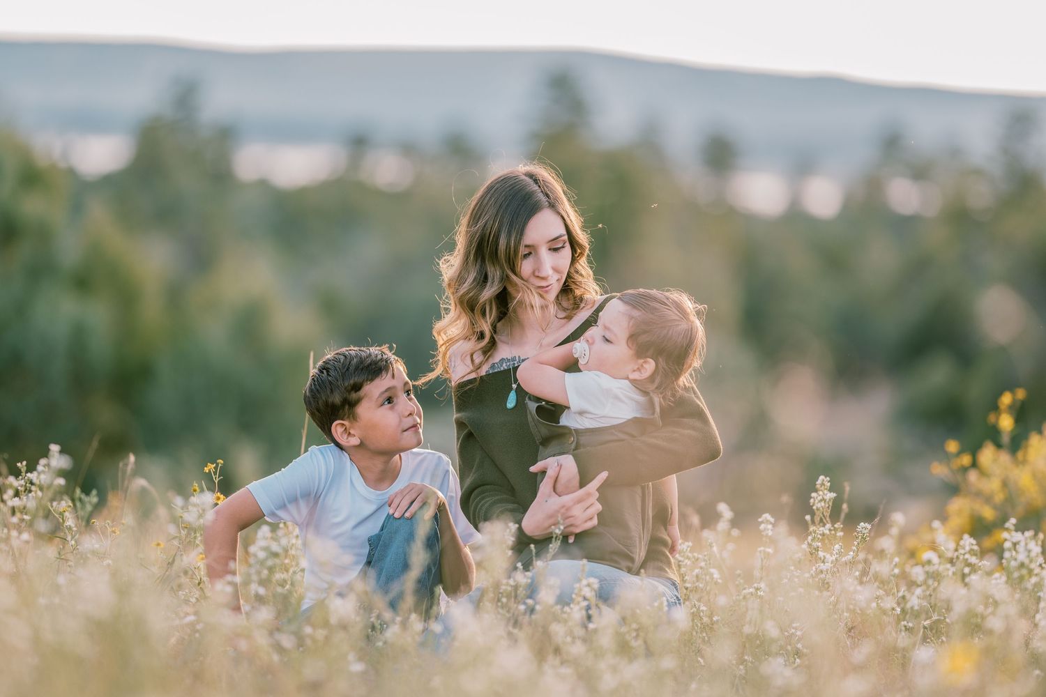 Mother with two toddlers sitting on green grass in soft evening light — tender family moment by Quiet Light by The Amoras.