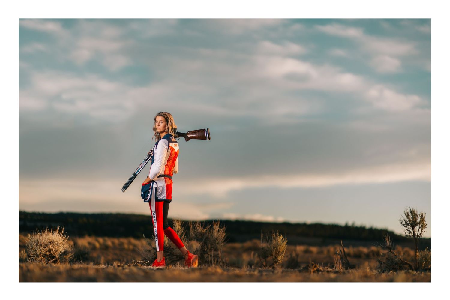 A young woman walks through an open field carrying a shotgun, wearing an orange hunting vest and red pants at dusk.