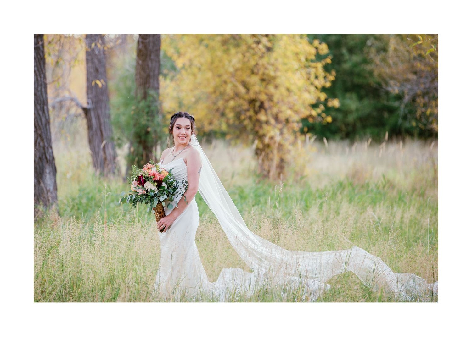 A wedding dress with a flowing white veil and bouquet of flowers stands gracefully in a sunlit meadow with autumn trees.
