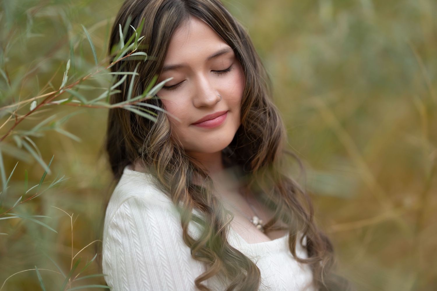 A woman in a white sweater stands in nature surrounded by willow branches with a serene expression in soft focus photography.