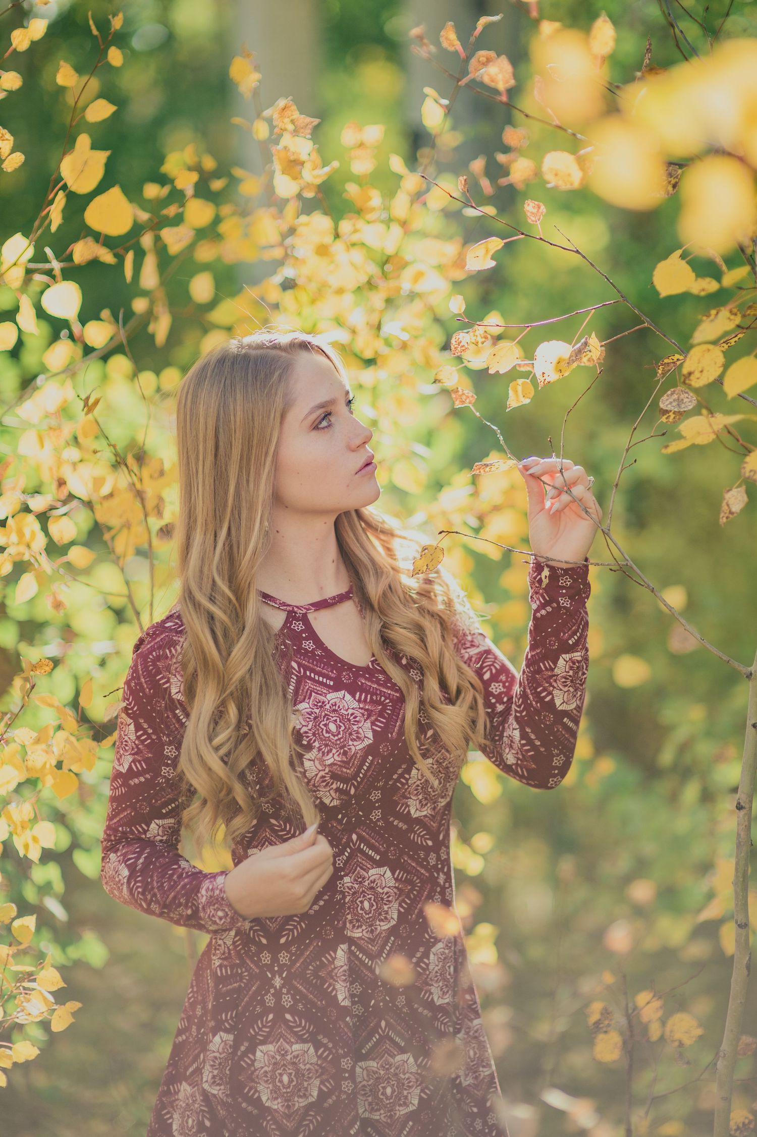Woman in maroon paisley dress standing among yellow autumn leaves in dreamy outdoor setting.