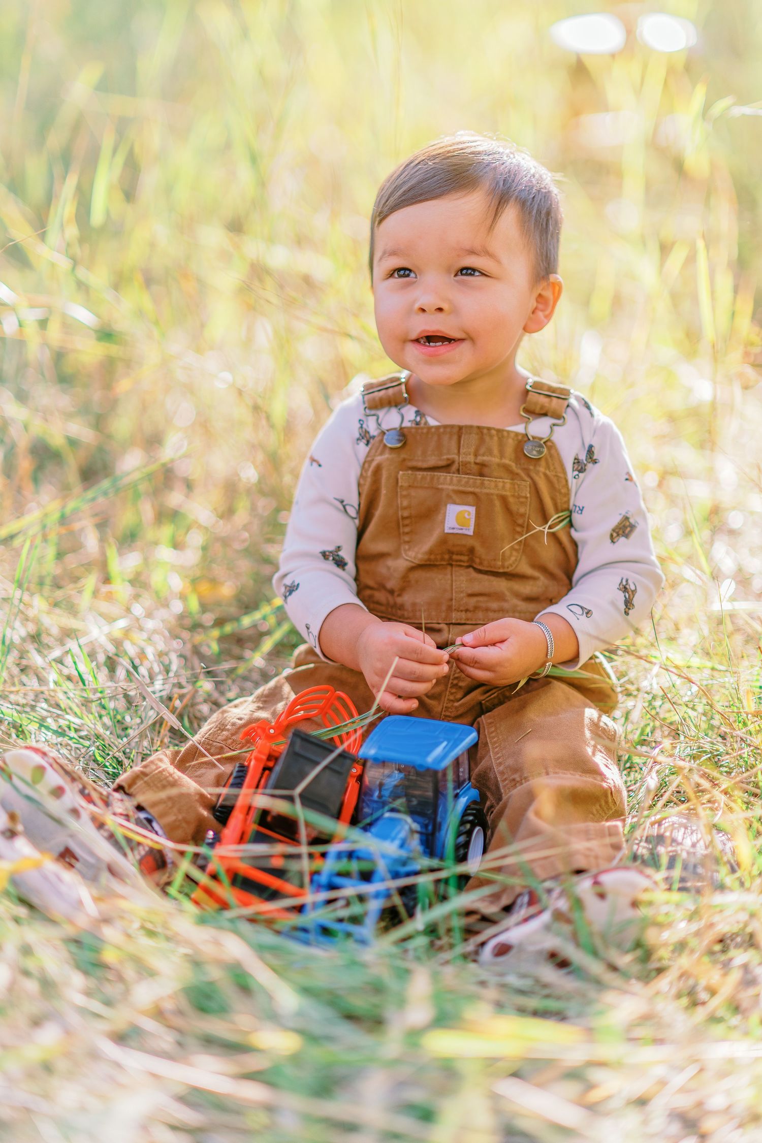 Baby in brown overalls sits in sunlit grass playing with colorful toy cars on a warm autumn day.