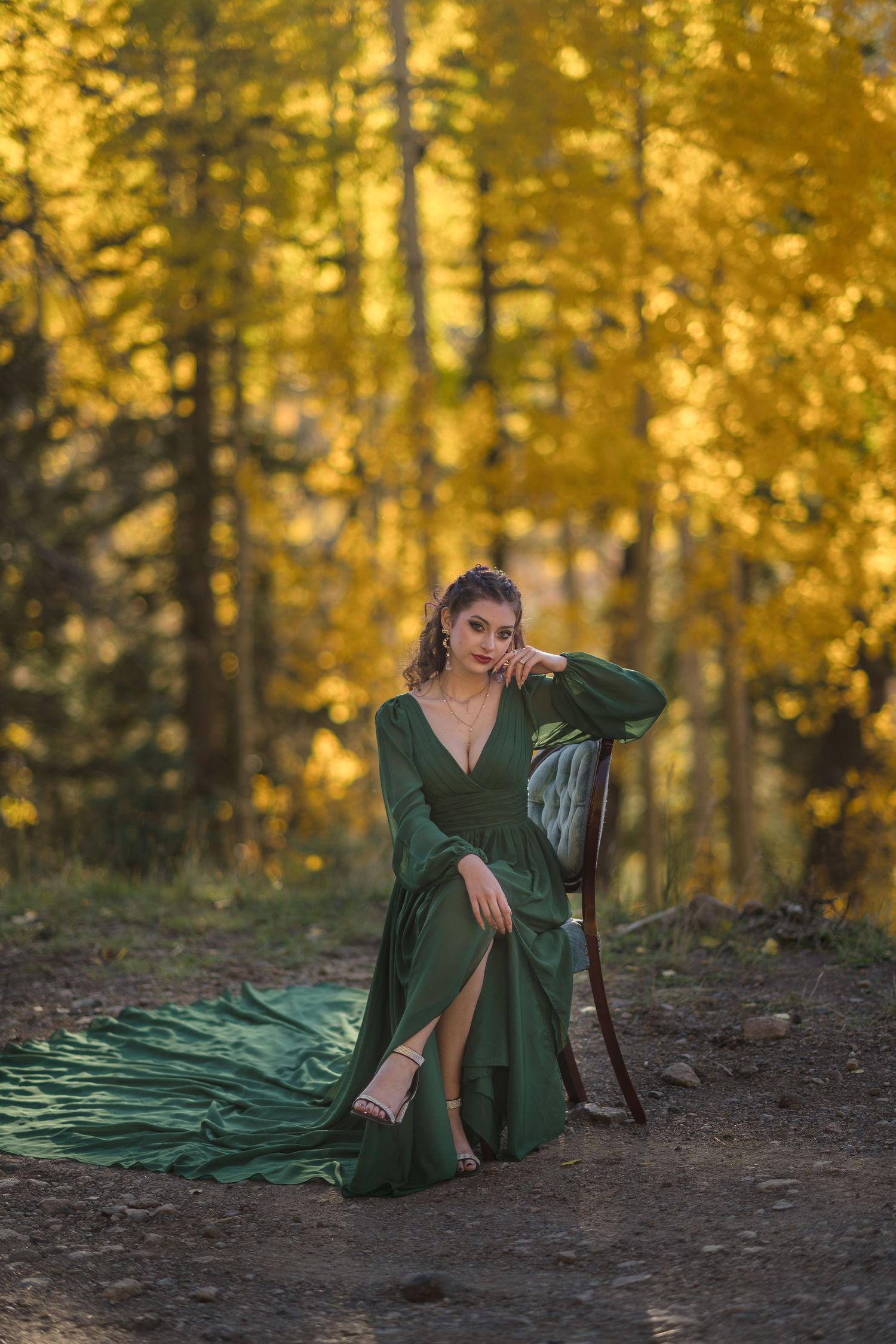 A figure in an emerald green dress poses gracefully on forest ground surrounded by golden autumn foliage.