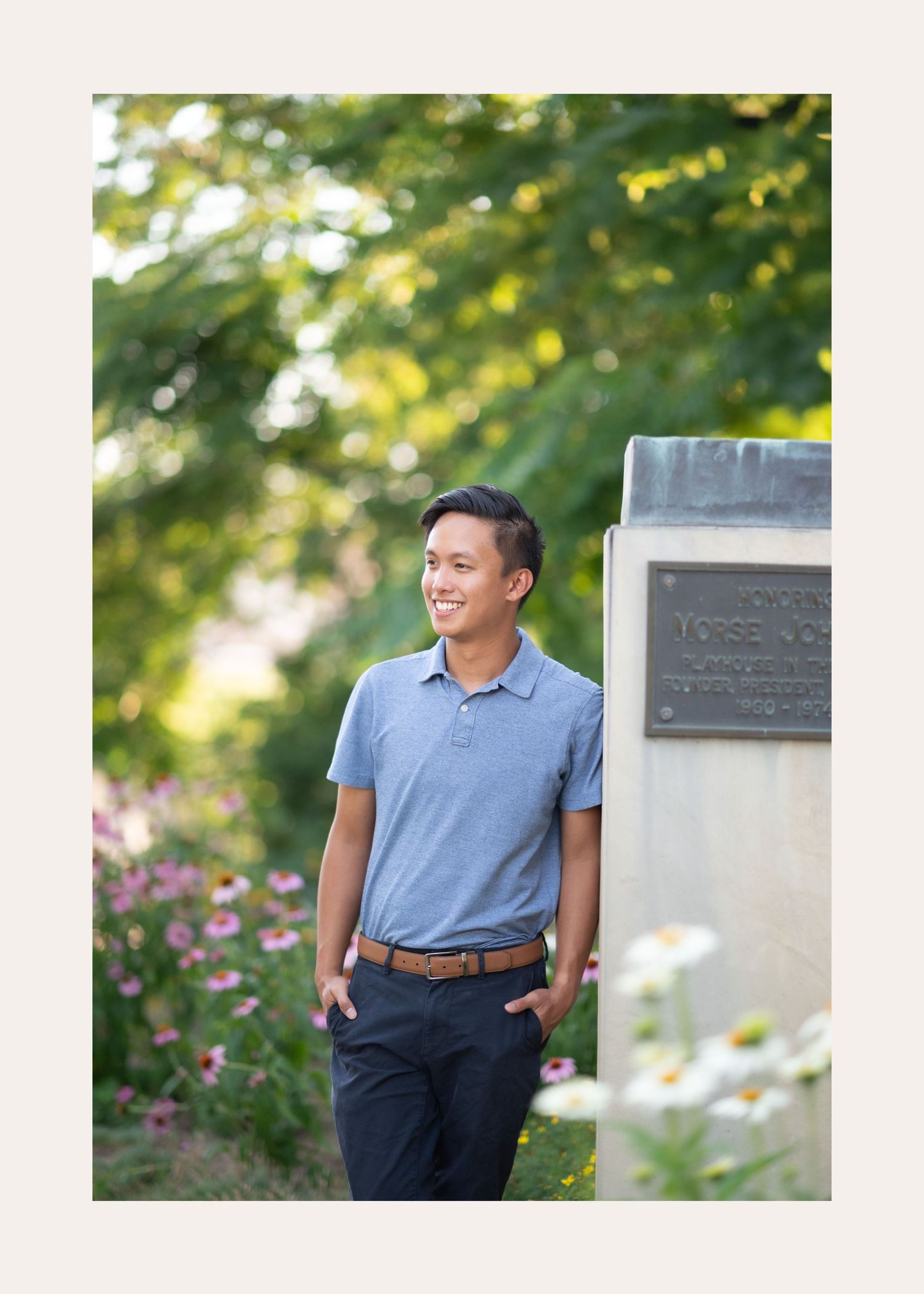 Someone in a blue polo shirt and dark pants poses casually against a stone monument in a garden with blooming flowers.