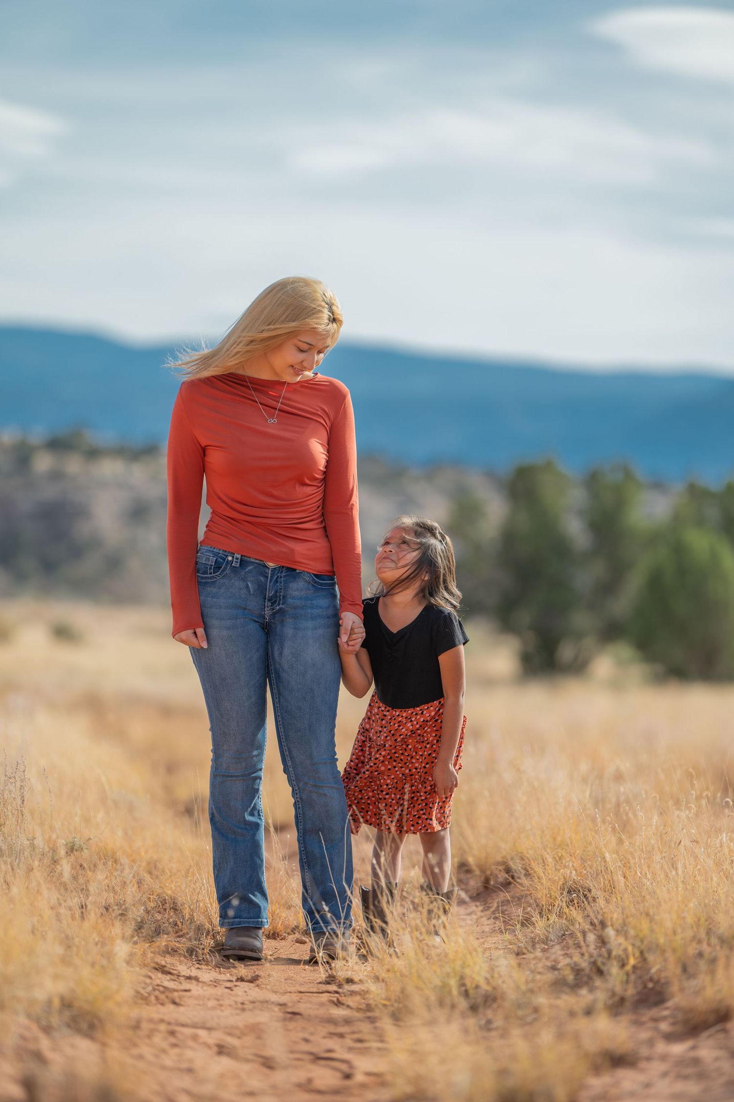 An adult and child walk hand in hand together through golden prairie grass with mountains visible in the distance.