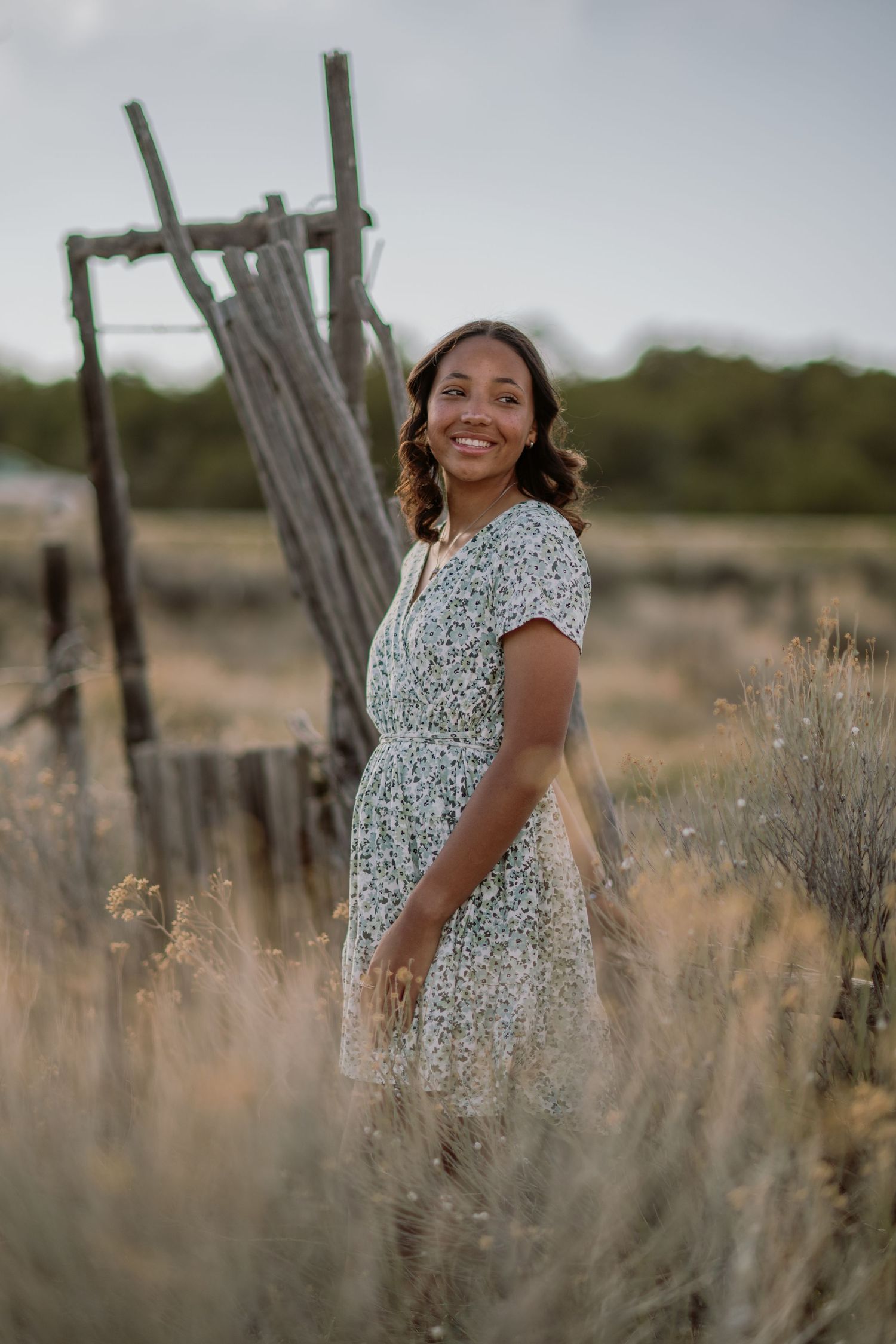 A person in a white floral dress poses cheerfully in a desert landscape with dried brush and wooden posts.