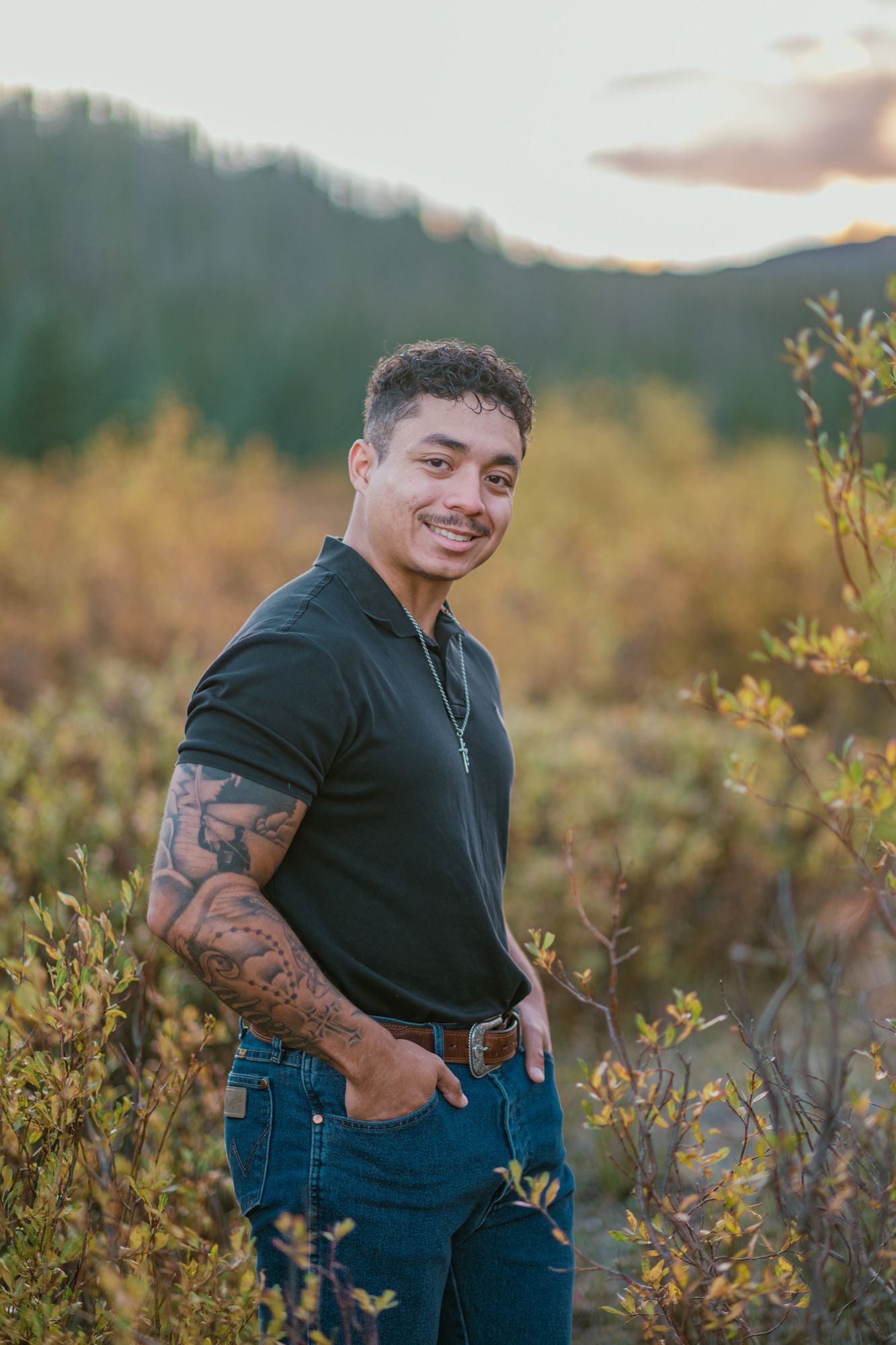Smiling young man with tattoos in black polo and jeans poses outdoors at sunset with mountain shrubs behind him.