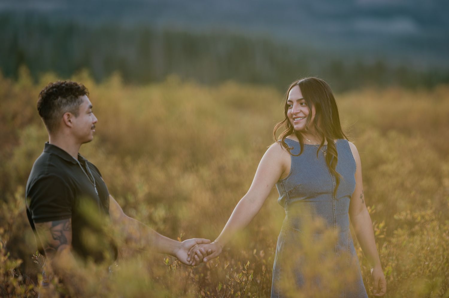 A couple holds hands and smiles at each other in a golden field during a romantic outdoor engagement photo session.
