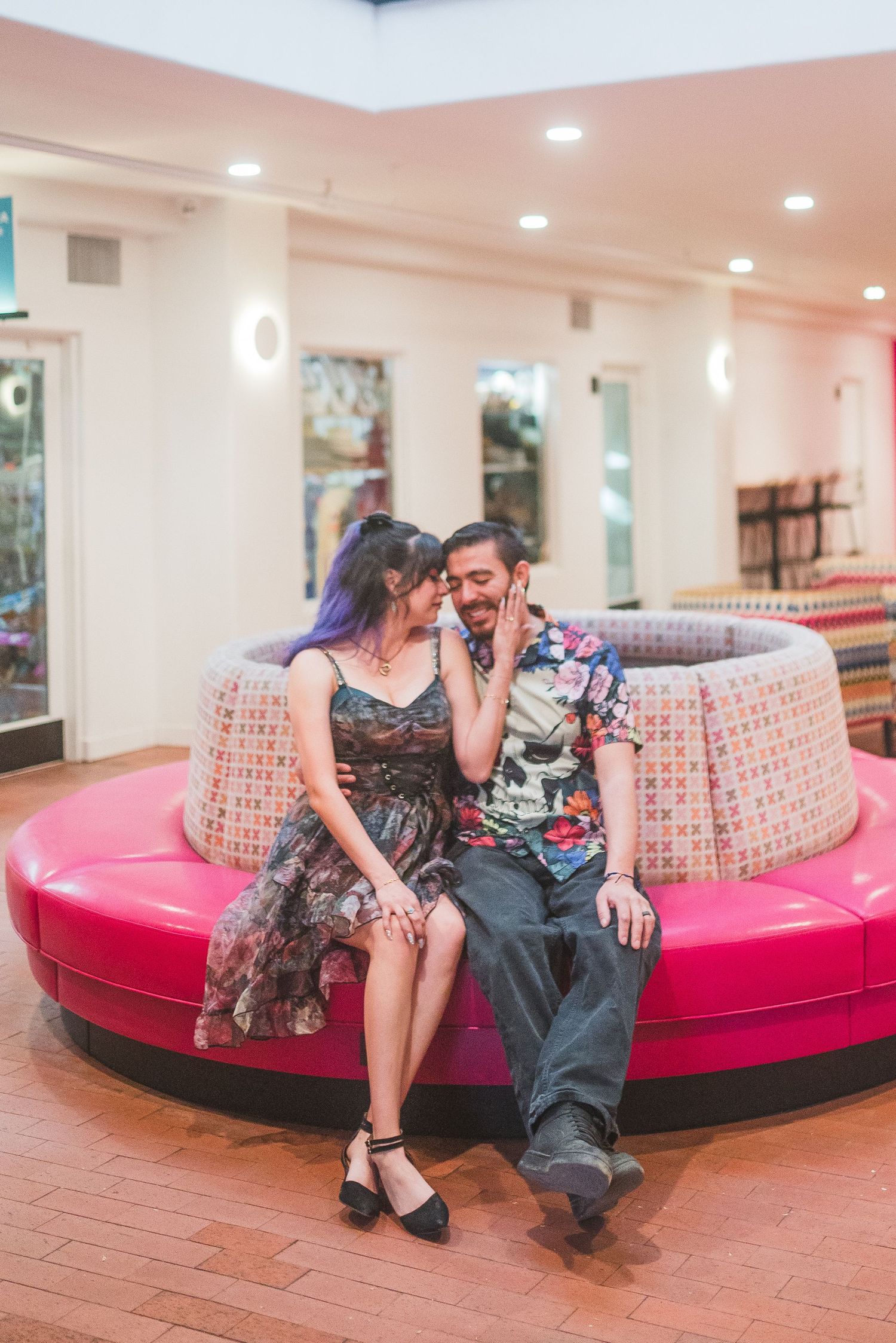 Couple sitting closely on a round pink bench in a colorful indoor venue, sharing an intimate moment.