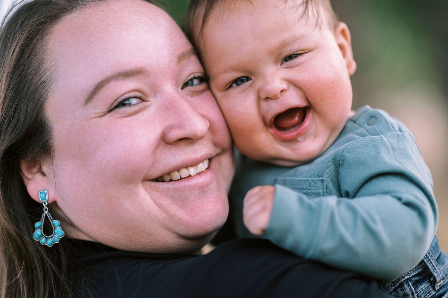 A smiling woman holds a laughing baby close to her cheek in a warm outdoor portrait.
