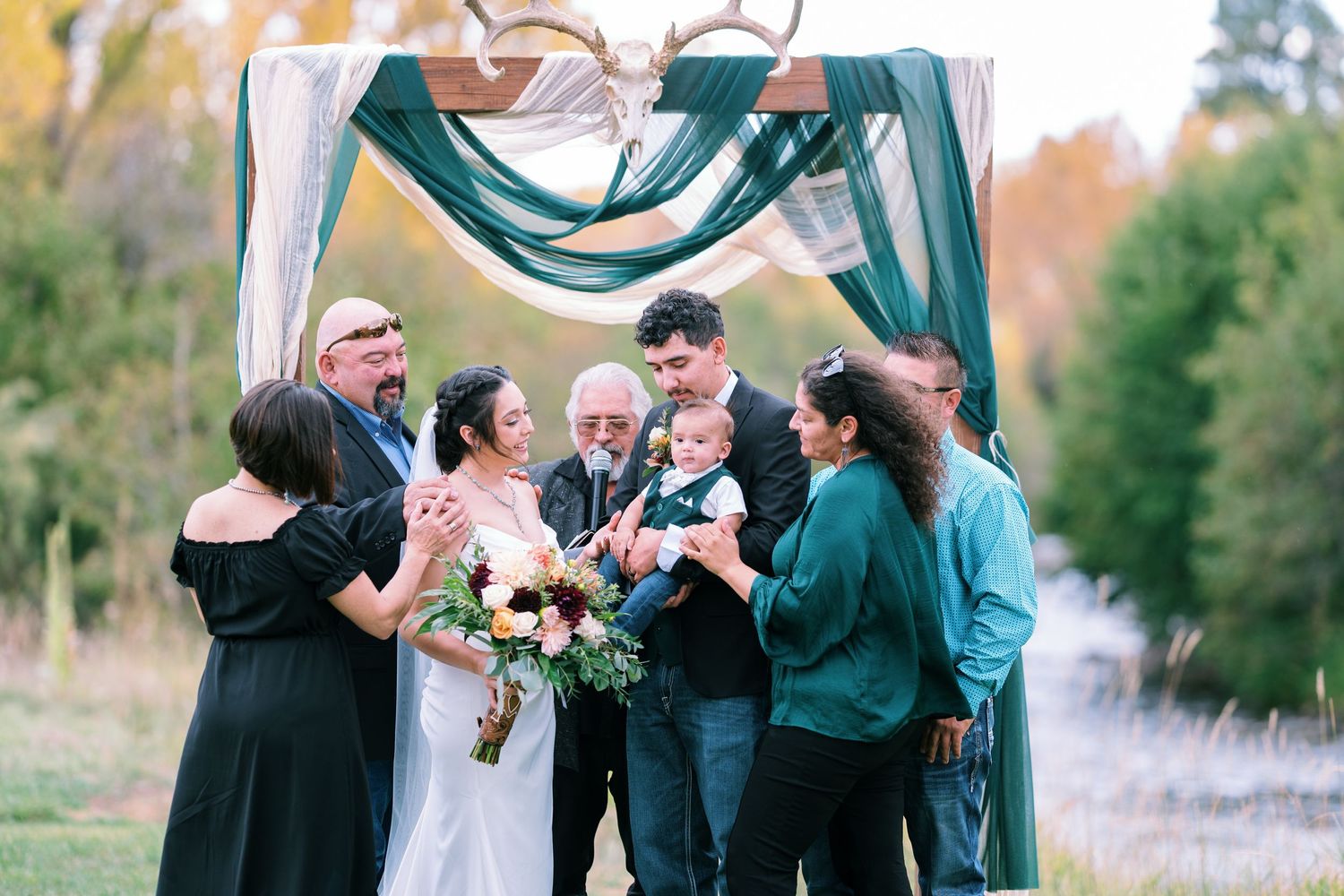 Wedding party gathered under teal and white draped arch sharing laughter during ceremony.
