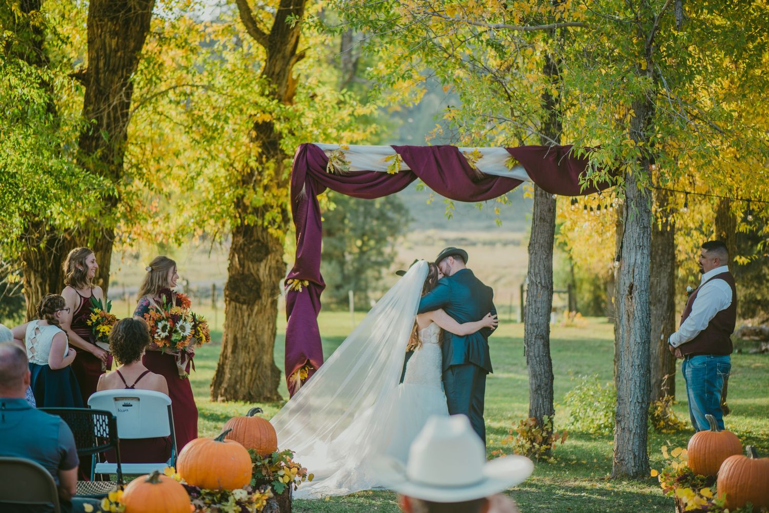 Fall wedding ceremony with pumpkin decorations under purple fabric arch and autumn foliage.
