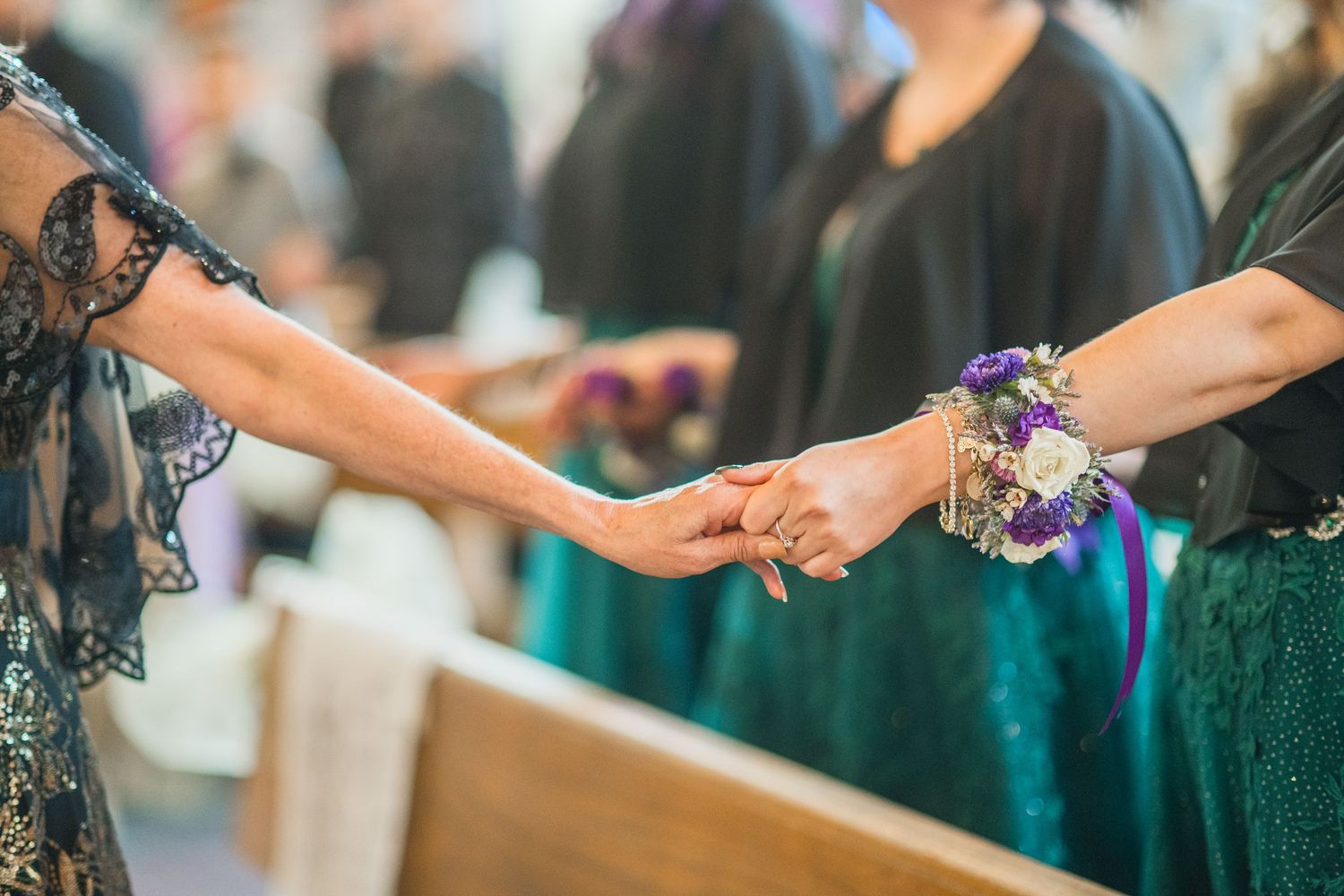 Two women holding hands, one wearing a floral wrist corsage with purple flowers at a formal event.