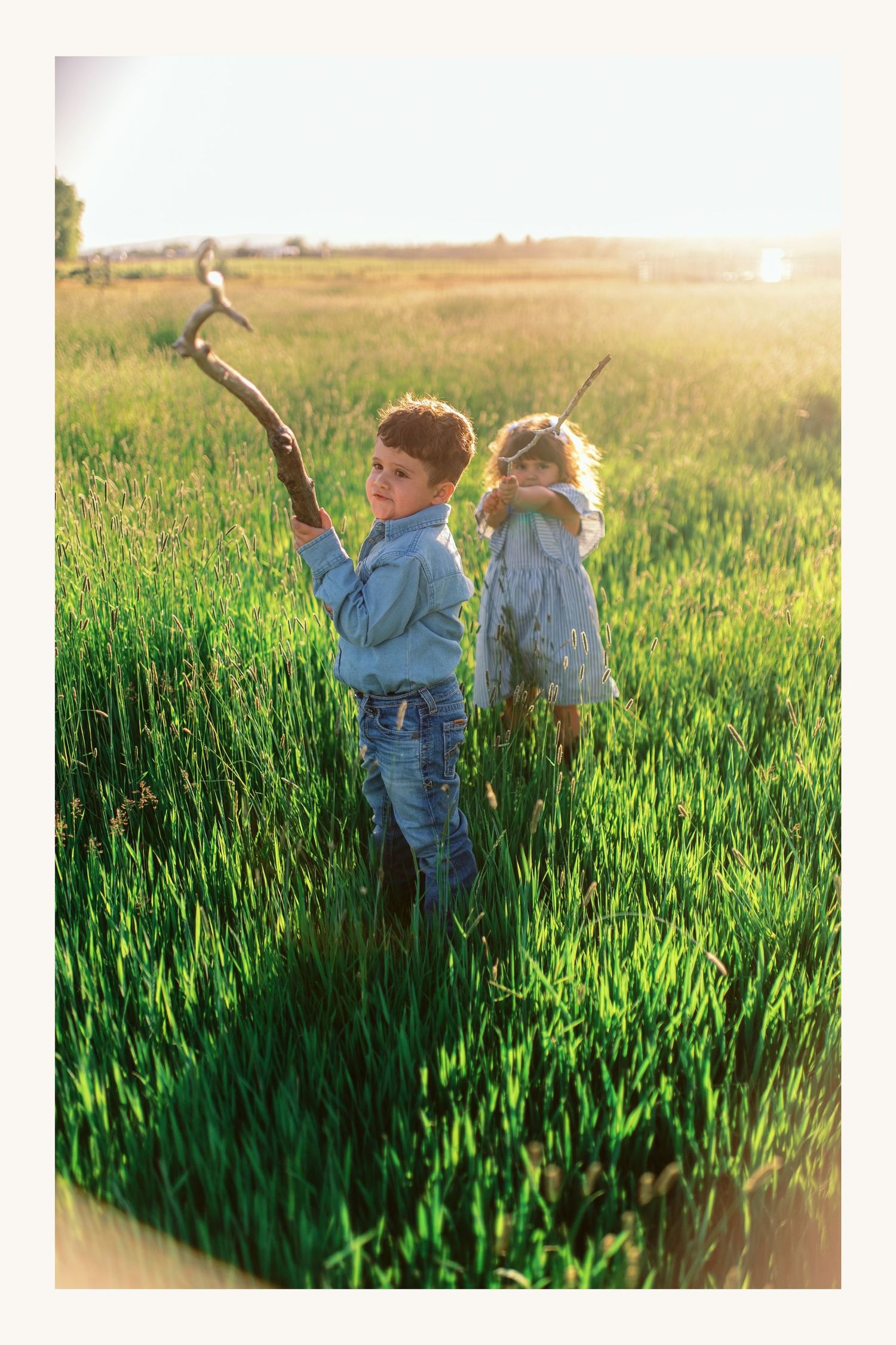 Two children playing in a sunlit green field, boy holding a stick, girl standing nearby, golden hour light.