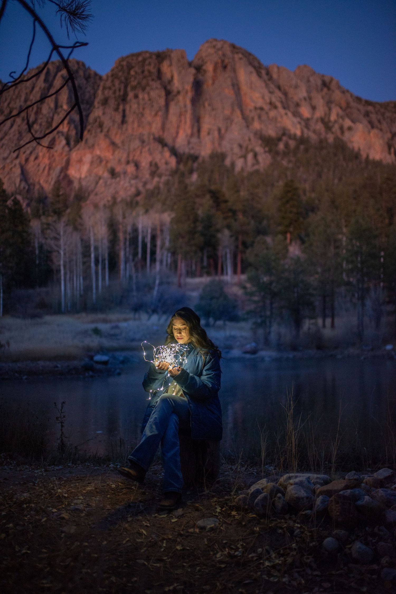 Woman holding glowing fairy lights by a mountain lake at dusk with red rocky peaks and forest backdrop.