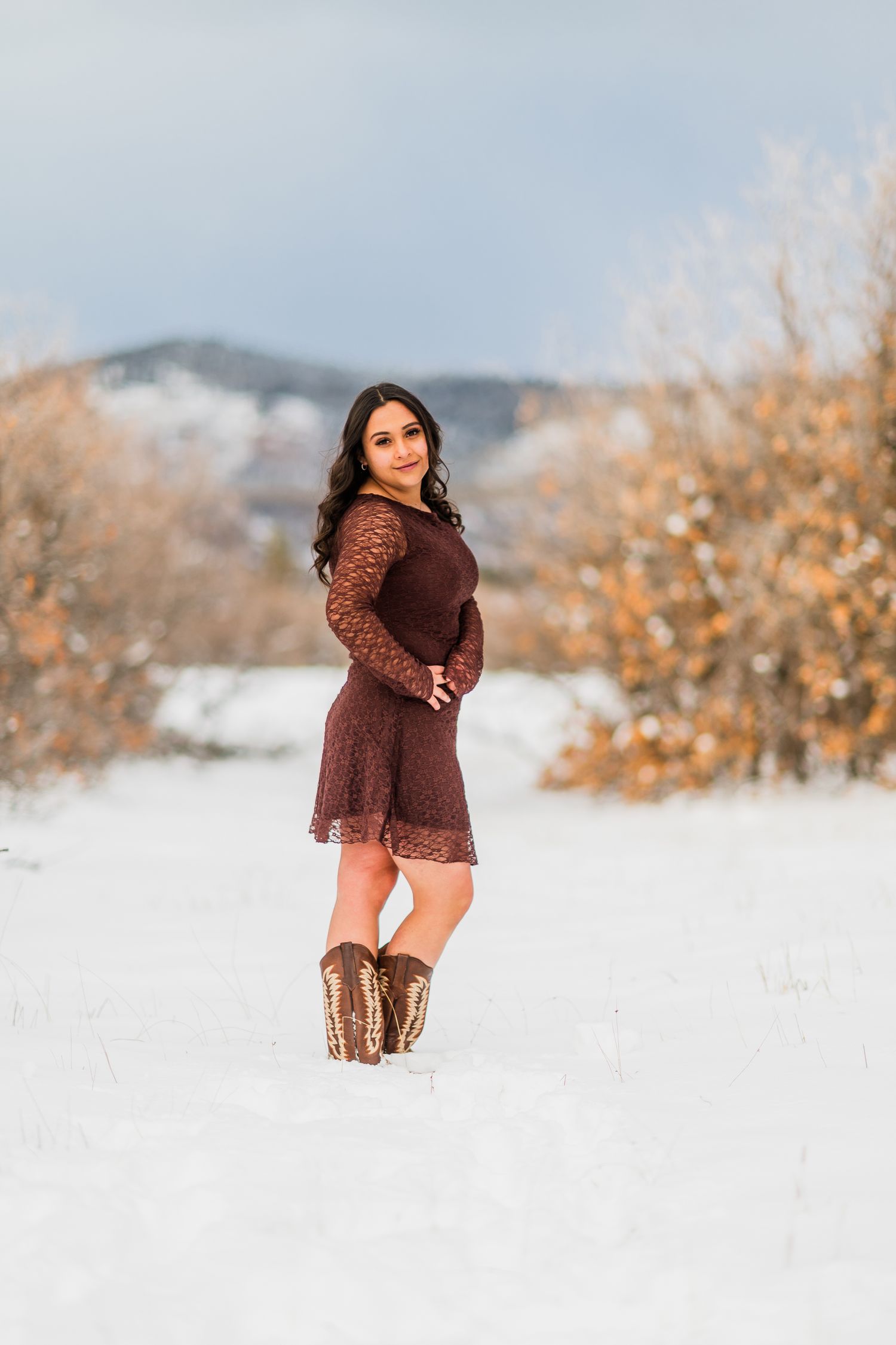 A person in a brown sweater poses in a snowy winter landscape with mountains and golden foliage in the background.
