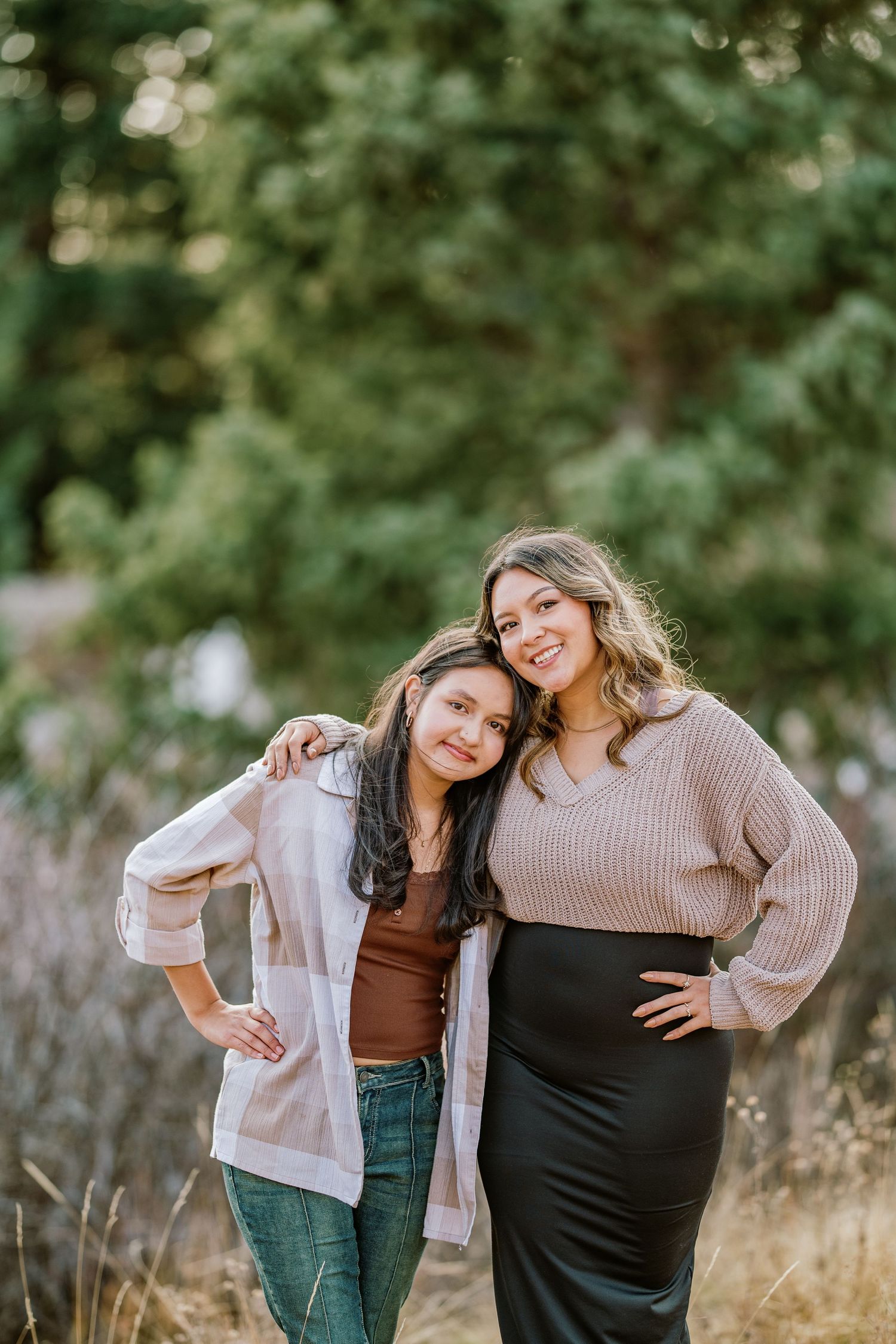 Two friends pose together outdoors wearing casual fall sweaters and standing in front of greenery in warm golden light.