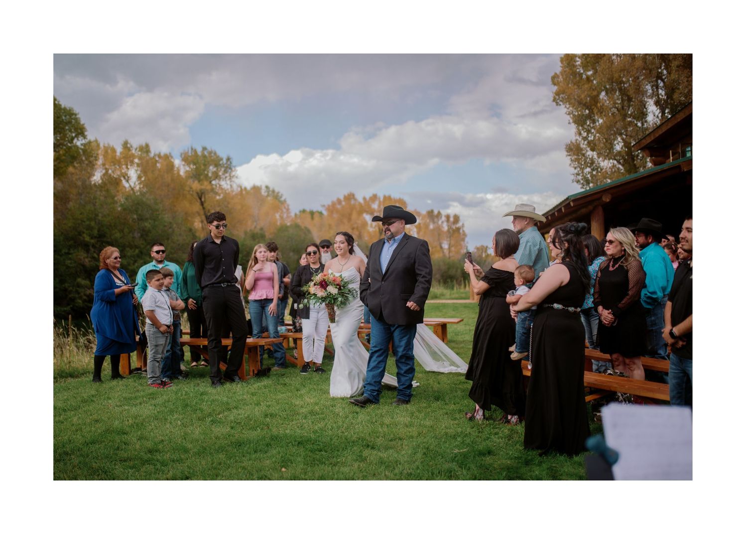 Outdoor wedding ceremony with guests arranged in a circle on grass with autumn foliage in background.