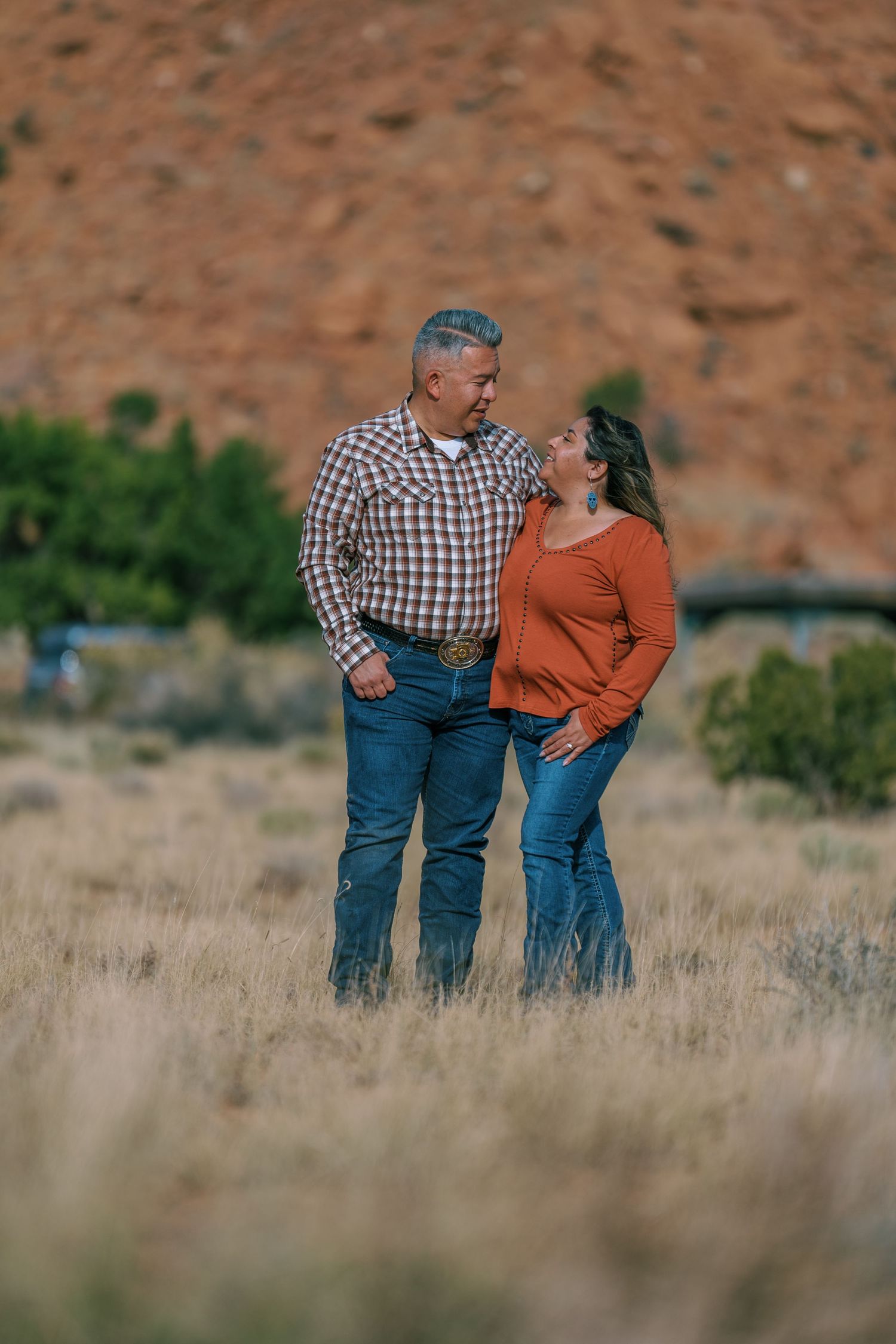 A couple stands together in a field of tall grass with red rock formations in the background.