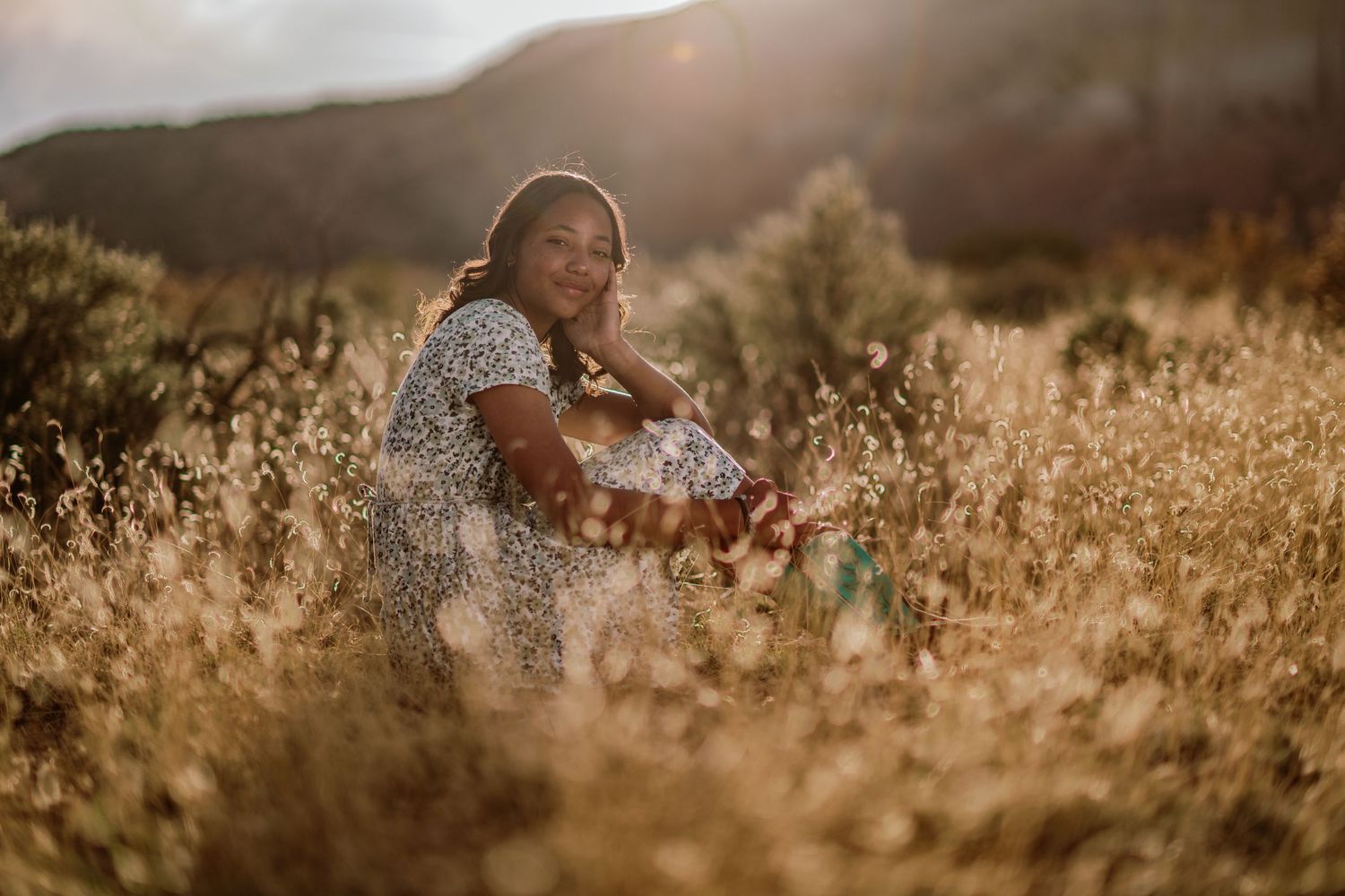 Someone sits in a golden field of tall grass at sunset with warm sunlight filtering through the mountains.