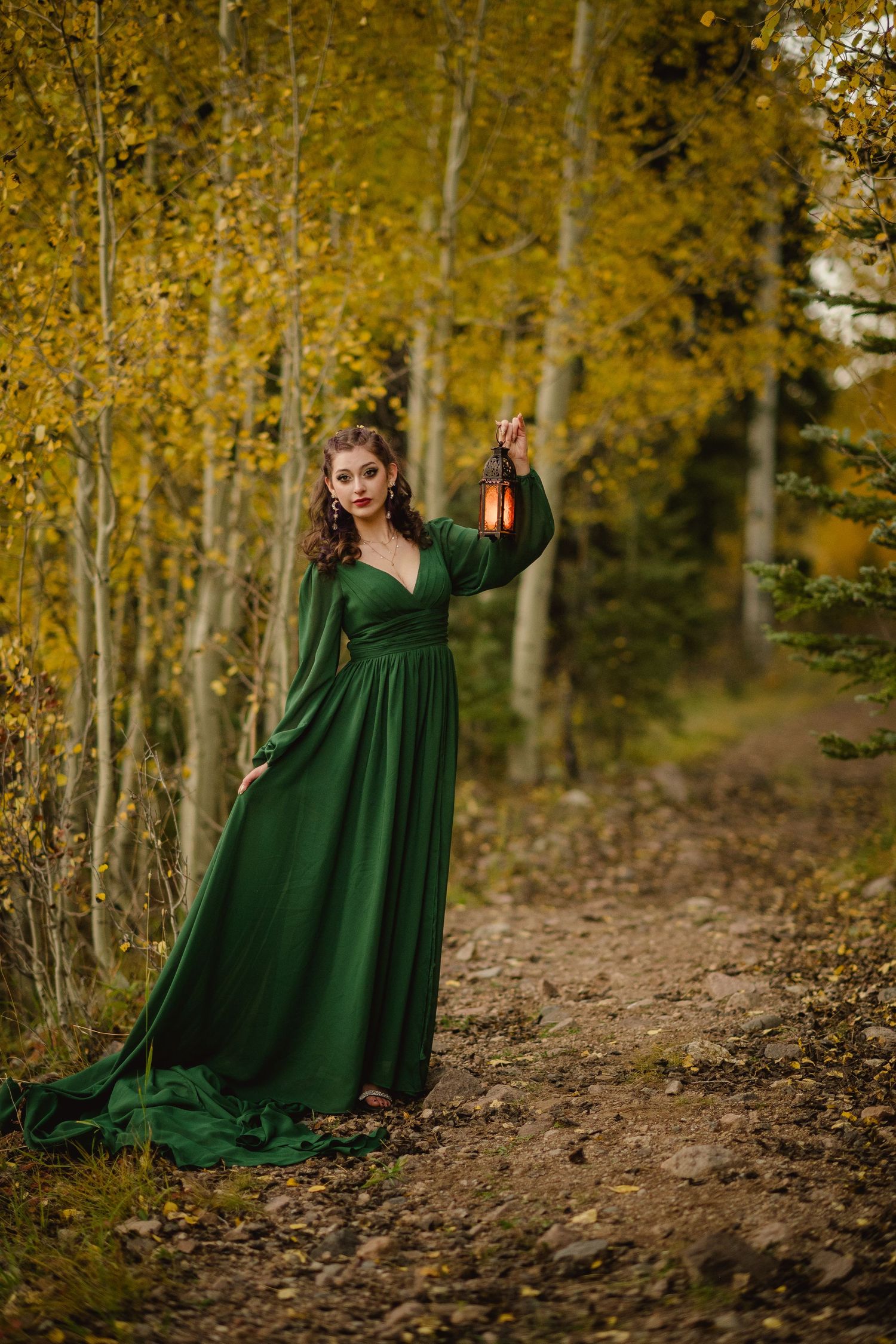 A woman in an emerald green dress poses gracefully among autumn birch trees on a leaf-covered forest path.