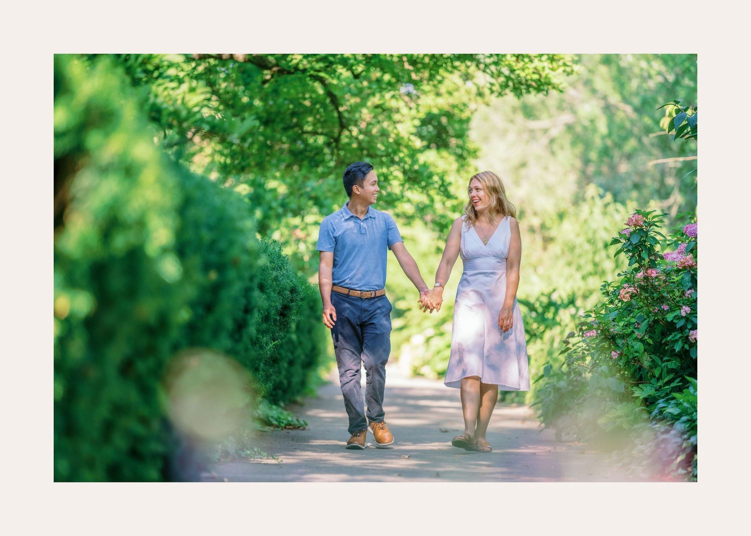 A couple holding hands while walking with their small dog along a lush green garden path in the sunshine.