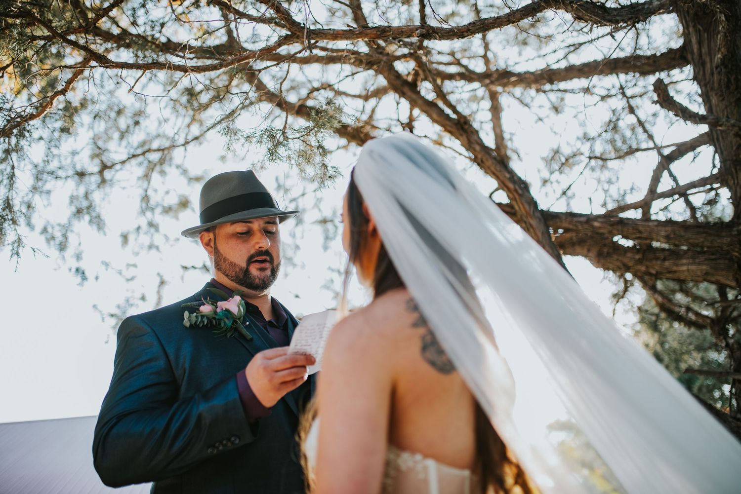 Wedding couple exchanges vows under a tree branch canopy during an outdoor ceremony.