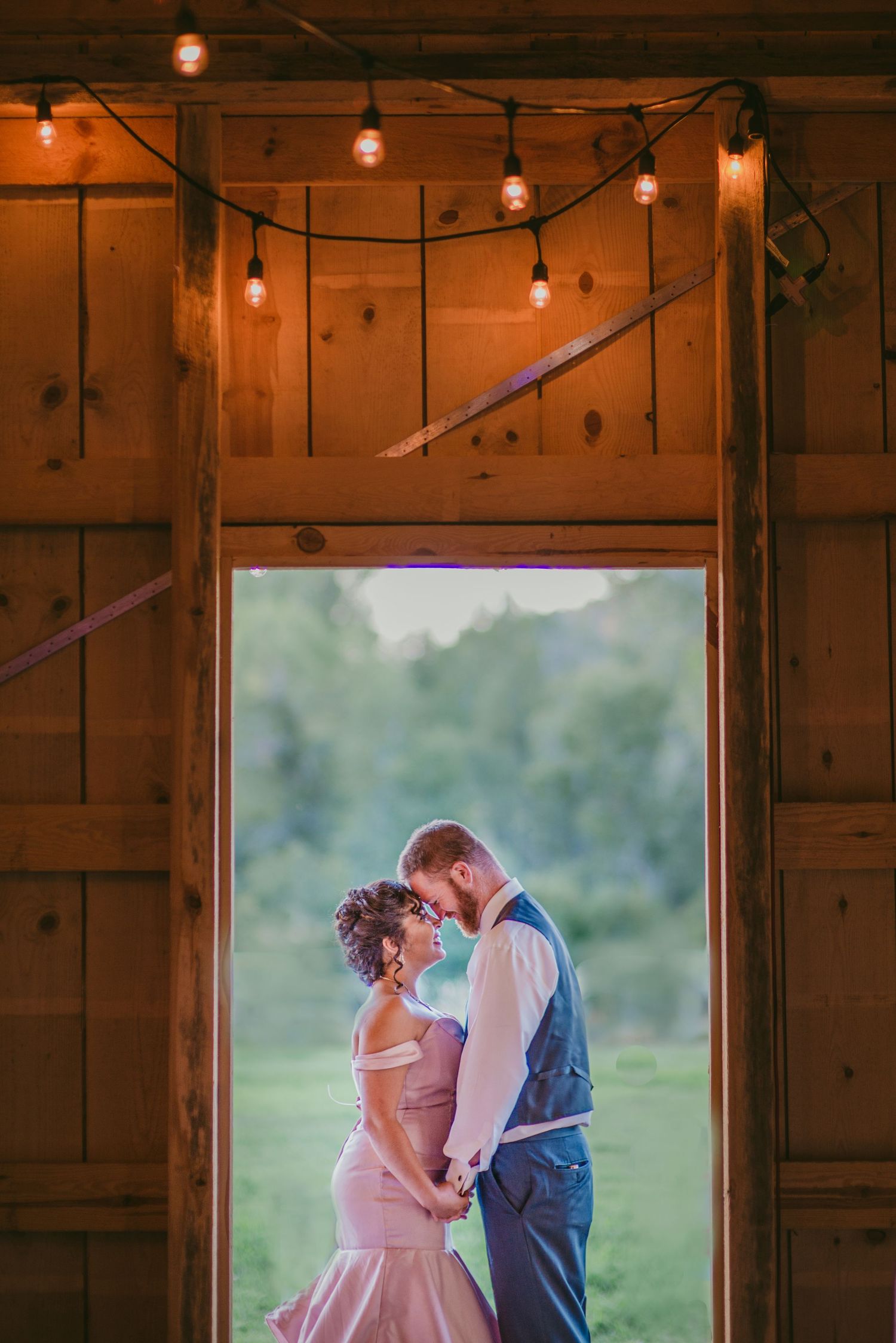 Romantic wedding scene in a rustic barn doorway with string lights overhead.