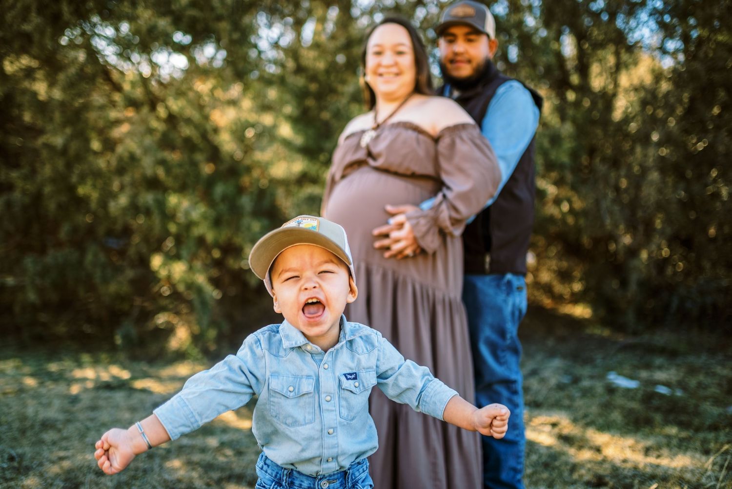 A joyful family photo session in a sunlit outdoor setting with natural greenery and trees in the background.