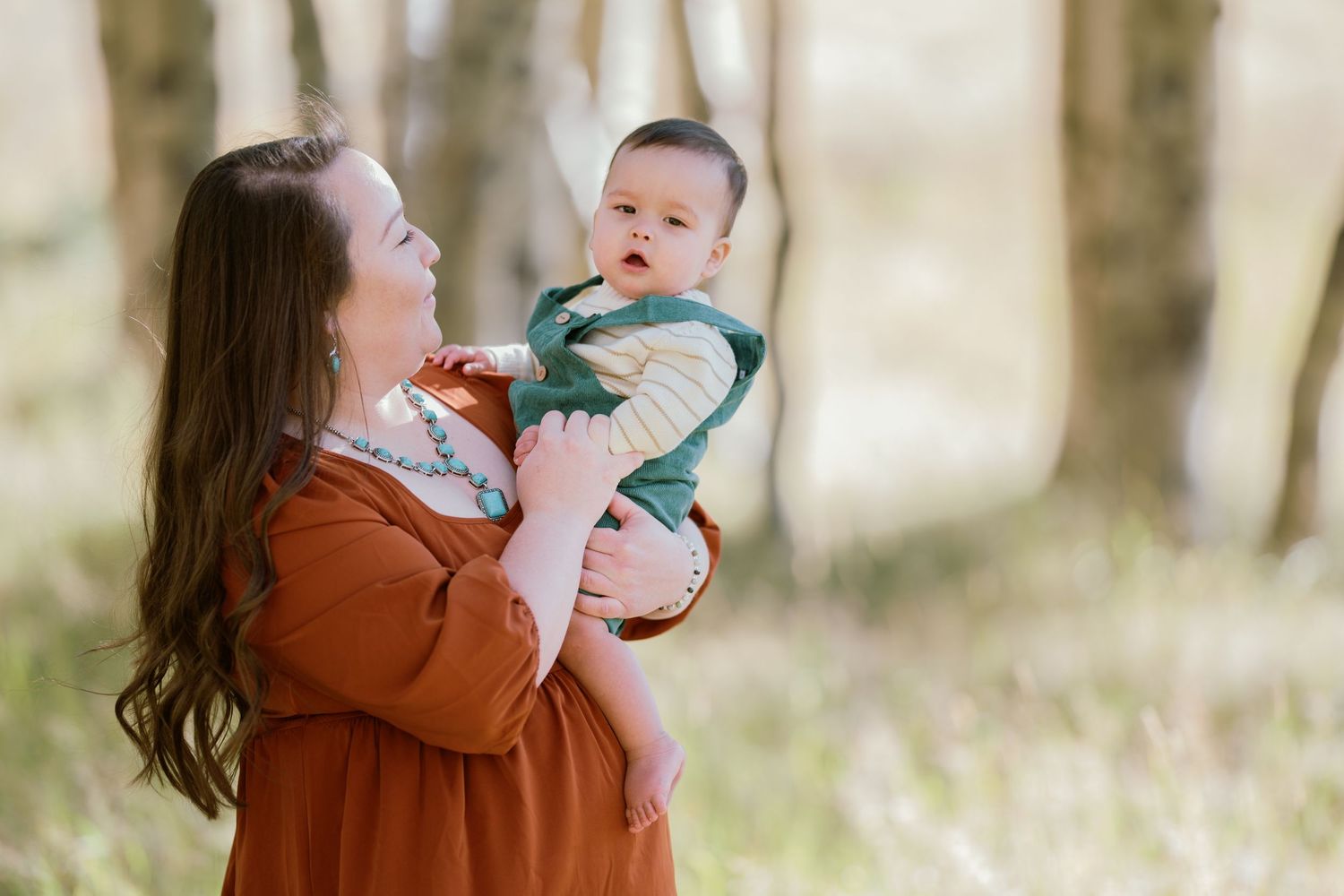 Woman in brown dress holding baby in woodland setting.
