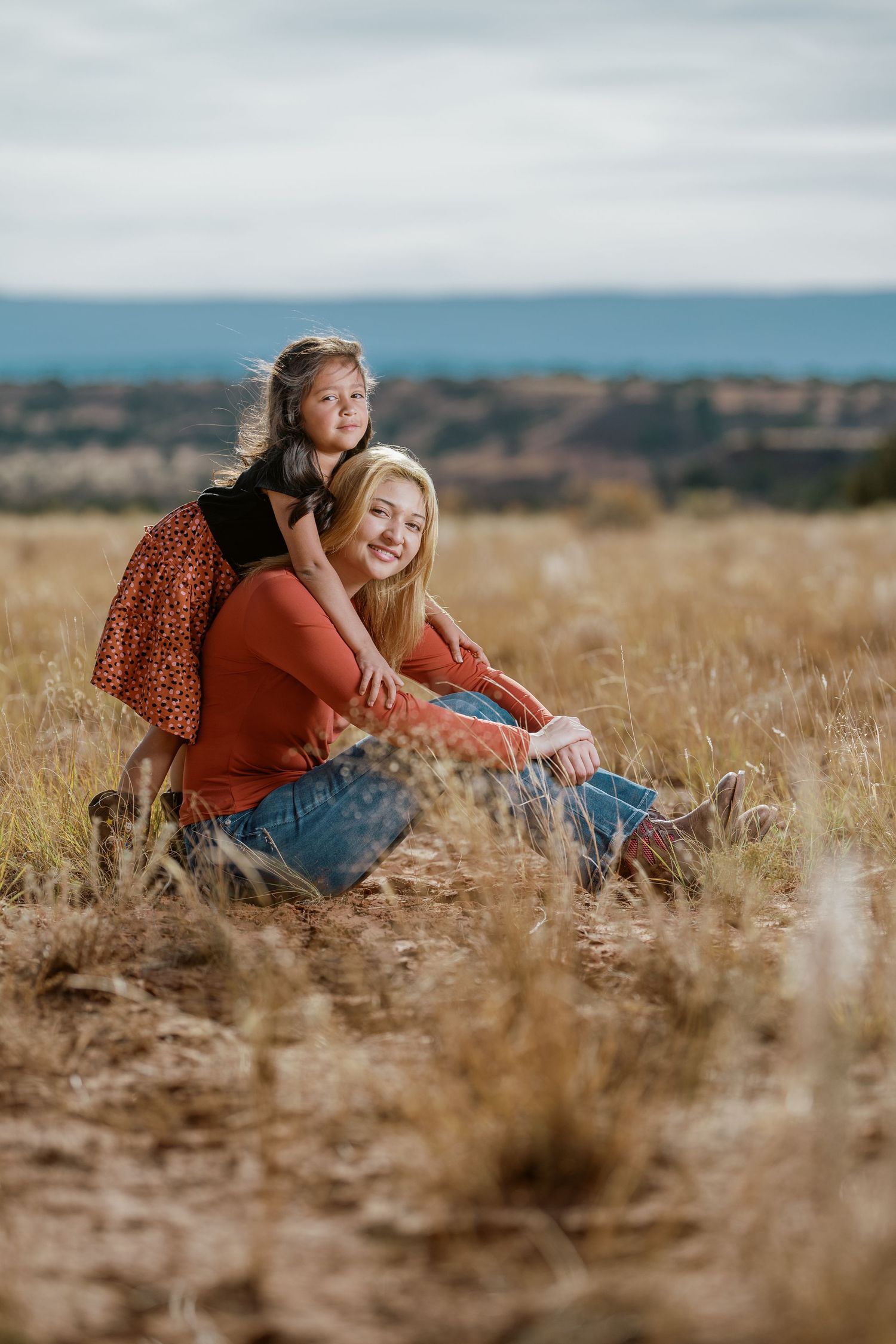 Two people share a candid moment while relaxing in a sun-drenched field with mountains in the background.