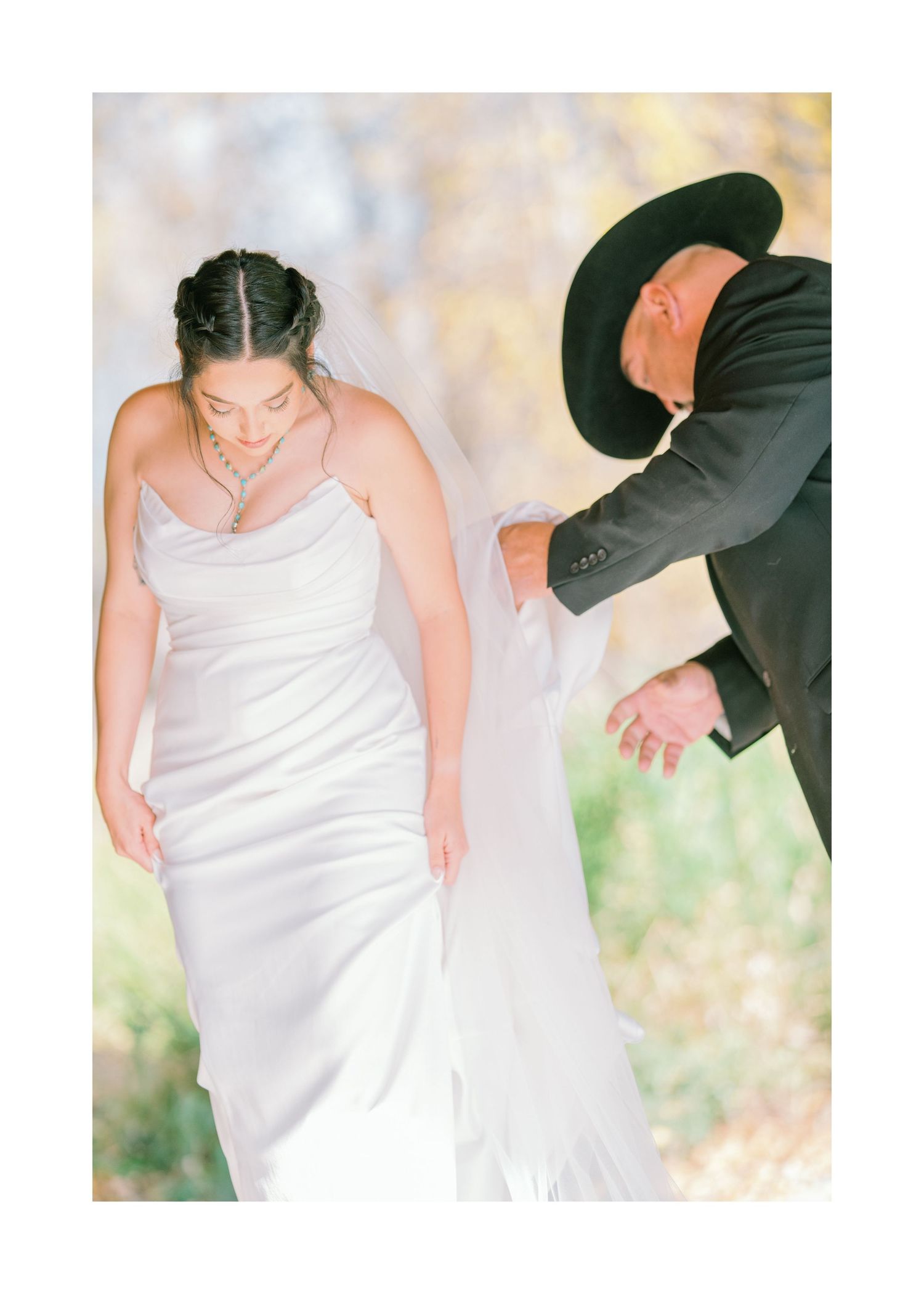 A bride in a sleek white wedding dress looks down while someone in a dark suit stands nearby in an outdoor setting.