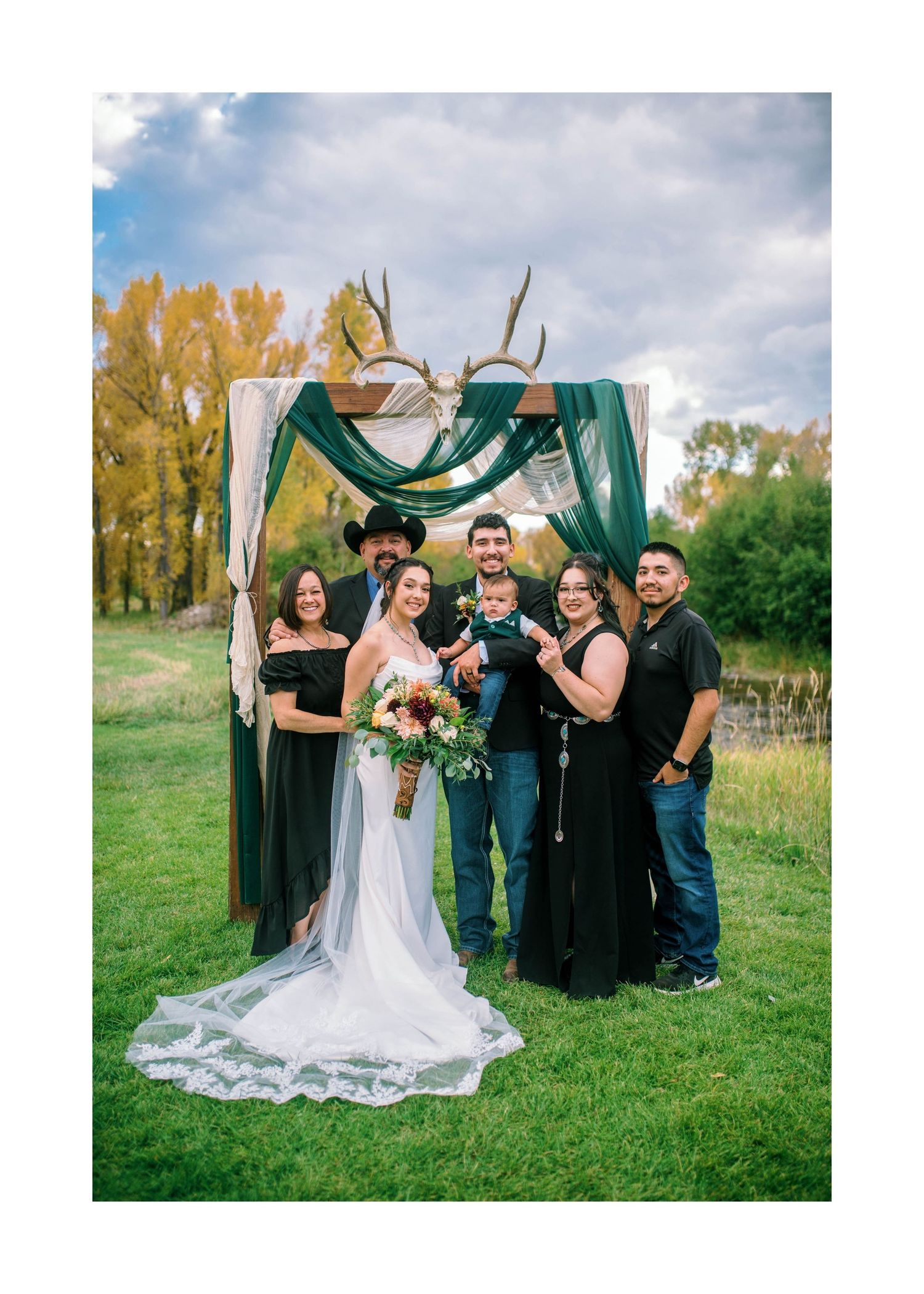 Wedding party celebrating outdoors with a decorative arch backdrop in a rustic outdoor setting with autumn trees and lawn.