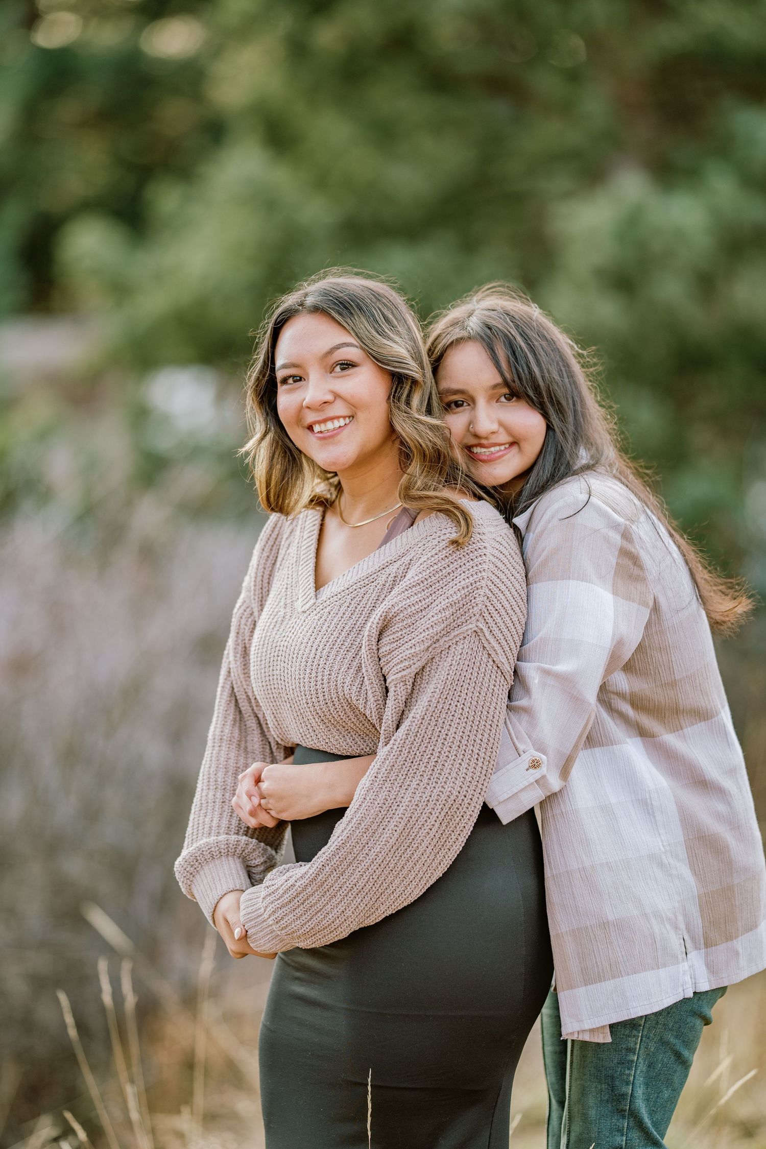 Two friends embrace and laugh together in a casual outdoor photoshoot with light filtering through trees.