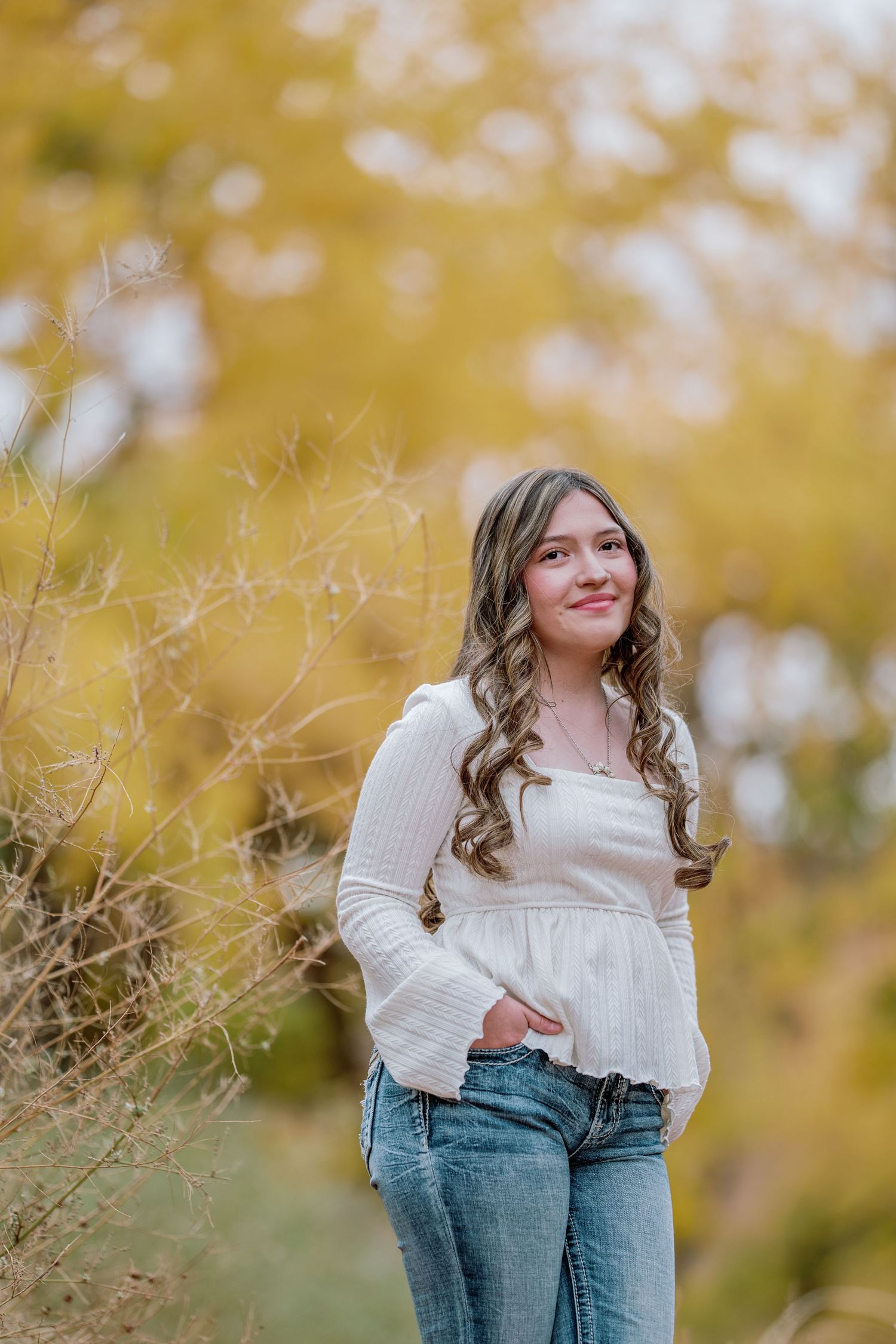 A person wearing a cream sweater and jeans poses outdoors against a blurred golden autumn background.