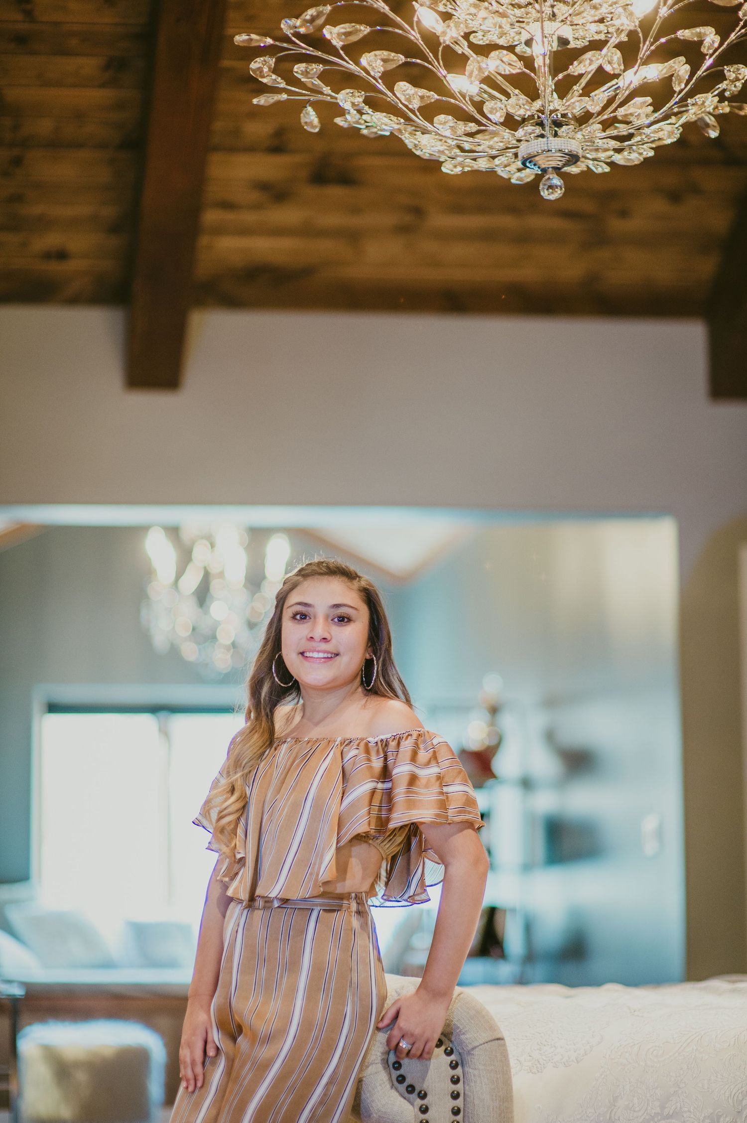 Senior portrait of a high school girl standing under a glowing chandelier — softly lit and elegantly captured by Quiet Light by The Amoras.