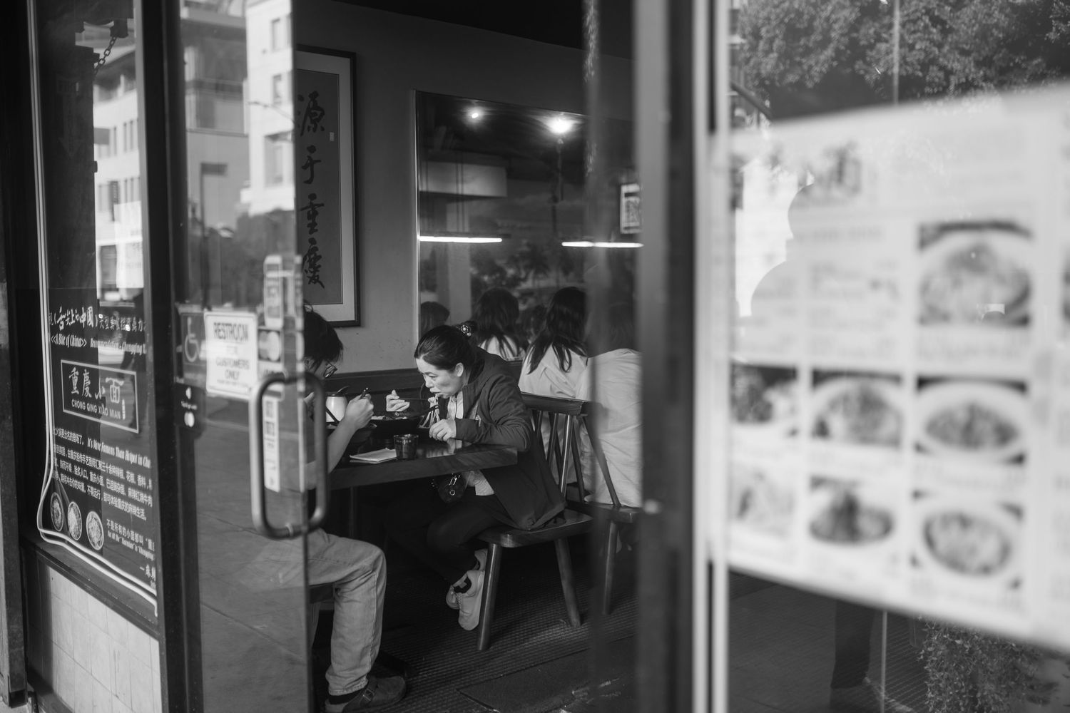 A black and white photograph shows glowing storefront signs and windows at night along an urban street.