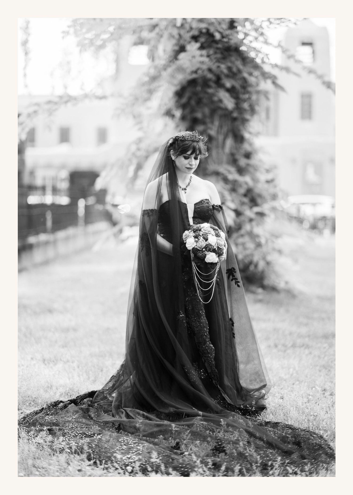 Black and white photo of a bride in a dramatic dark gown with long veil holding a bouquet outdoors.
