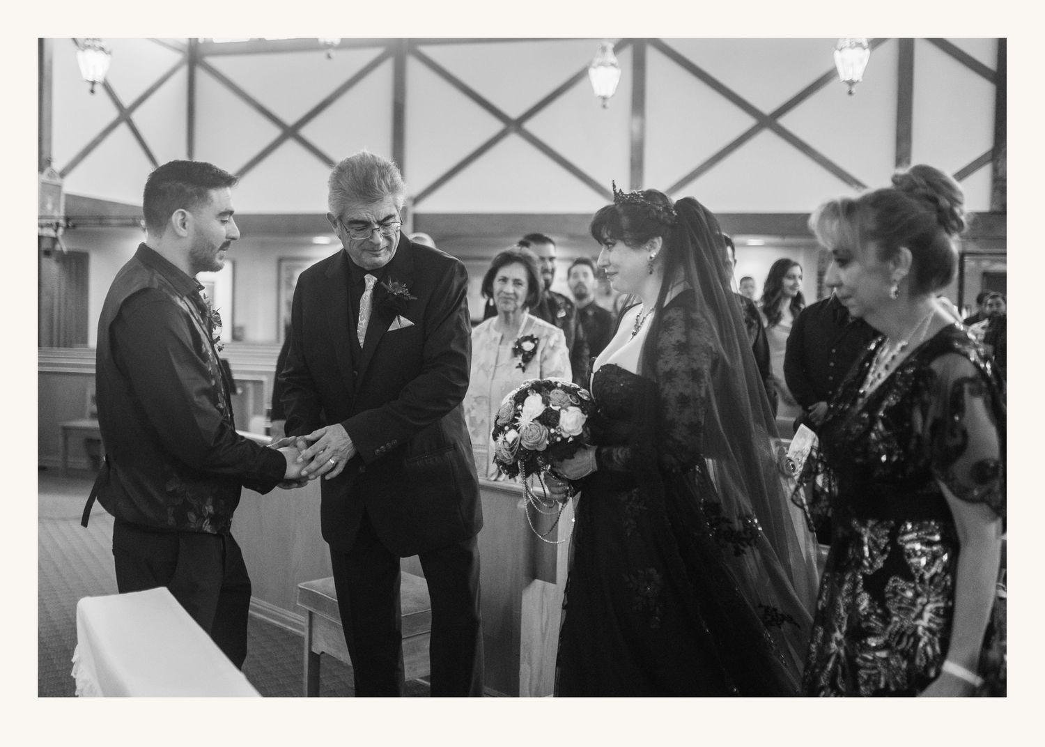Black and white wedding ceremony photo showing bride with bouquet, groom, and guests exchanging rings indoors.