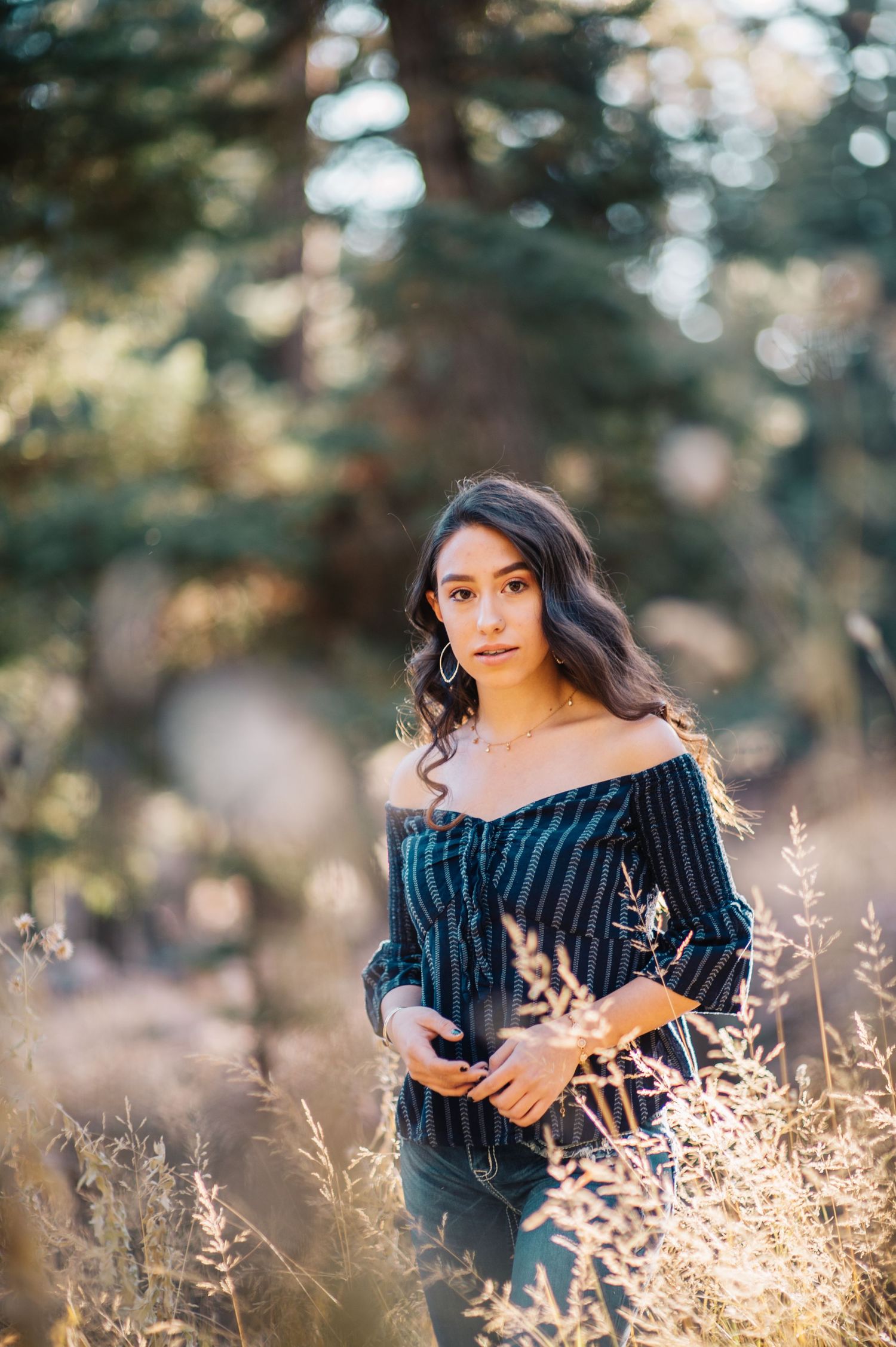 Young woman in striped off-shoulder top standing in sunlit field with tall grass and trees.