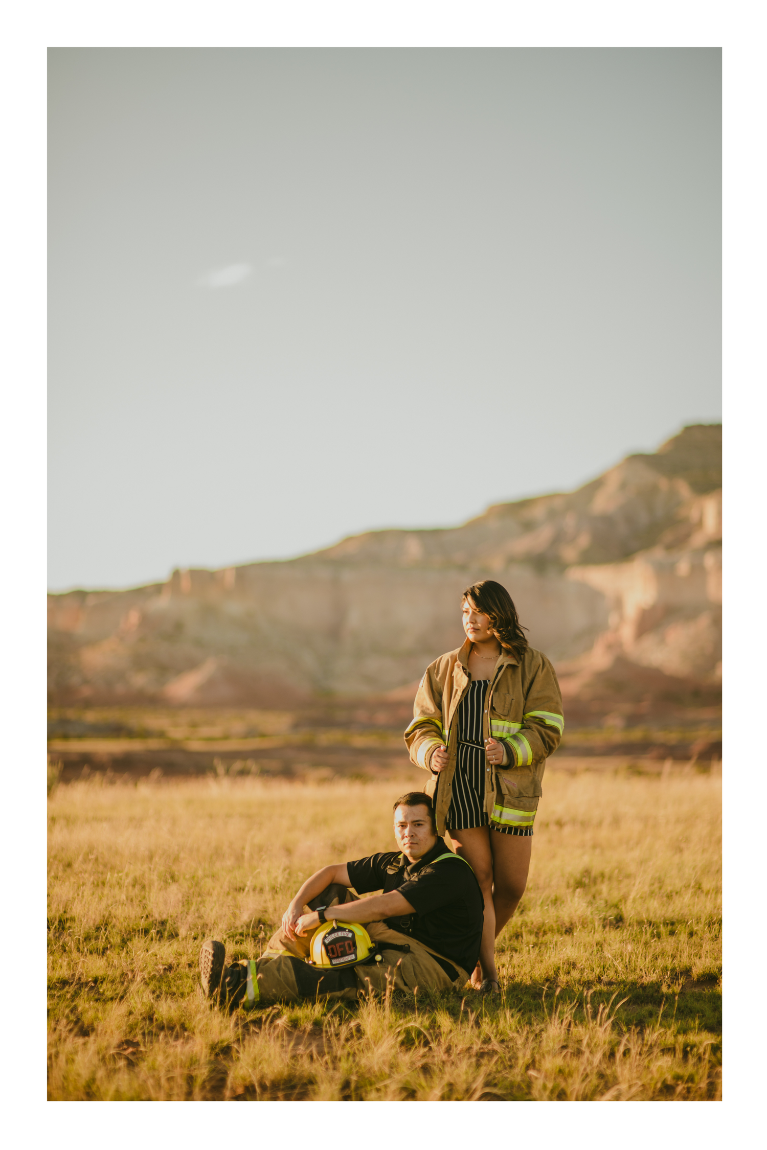 Couple in firefighter gear posing in golden field with red rock formations at sunset.