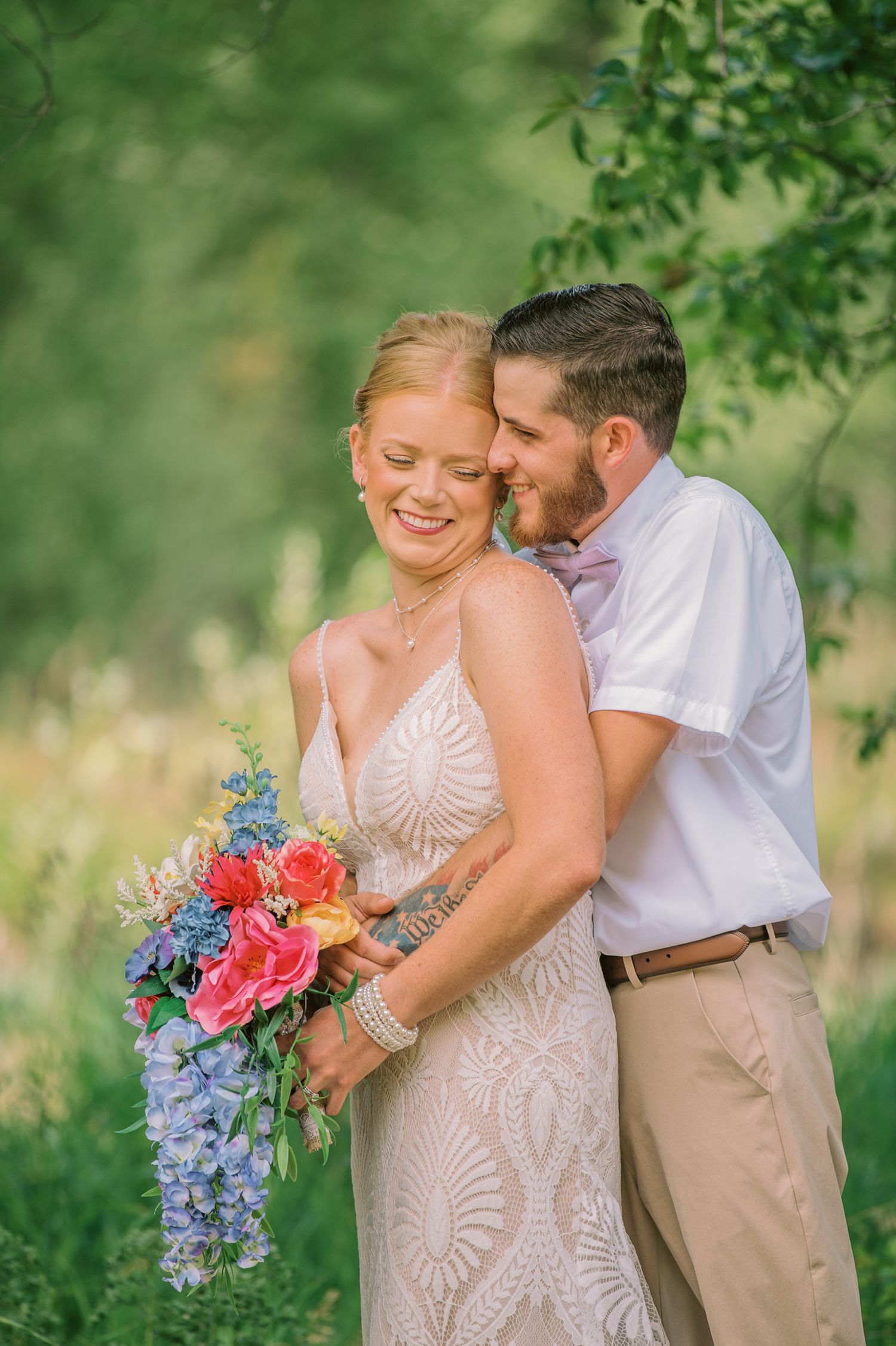 Bride in lace gown holds colorful bouquet as groom in white shirt embraces her in lush green outdoor setting.