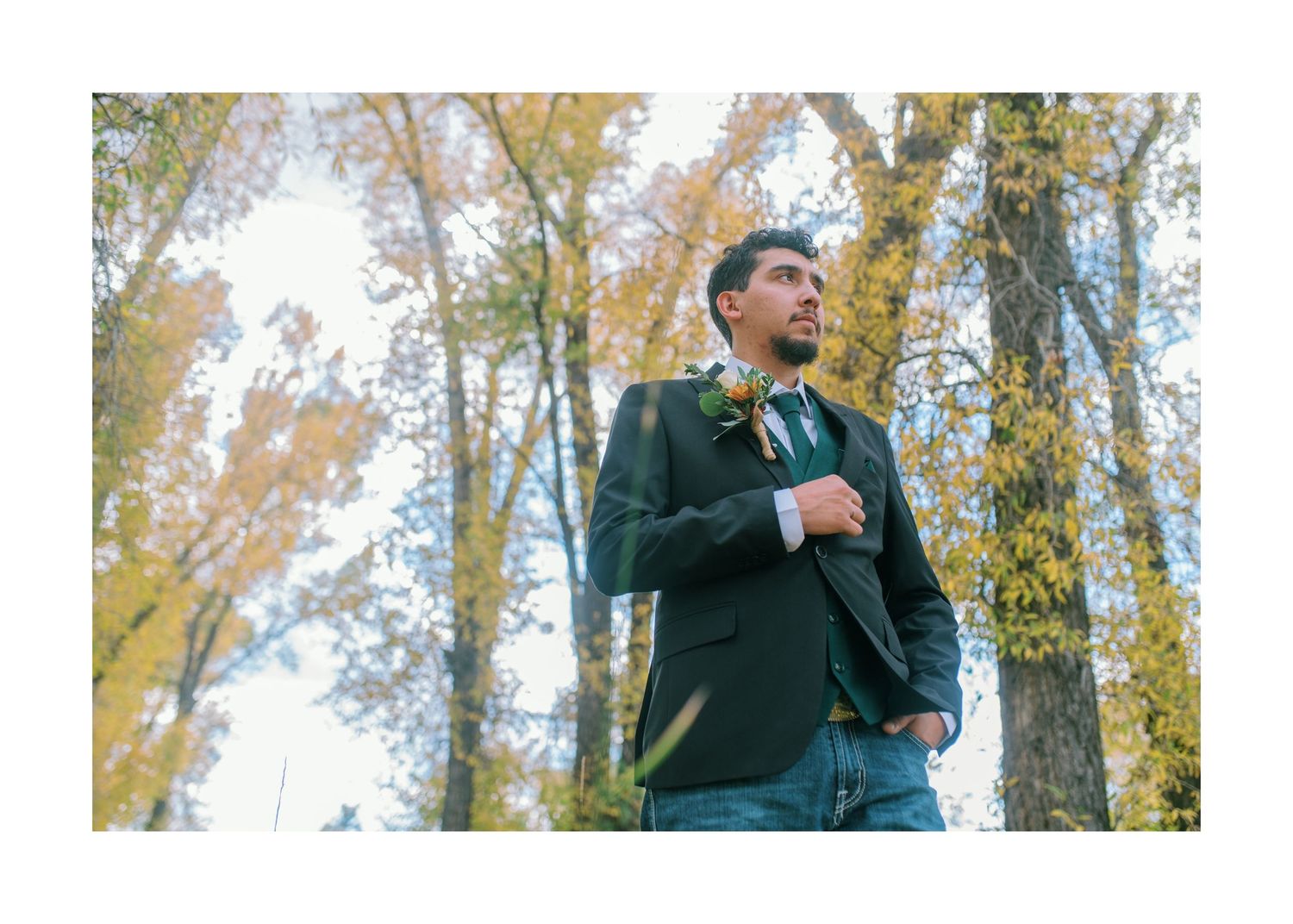 Someone wearing a dark green blazer poses in a wooded area with autumn trees in the background.