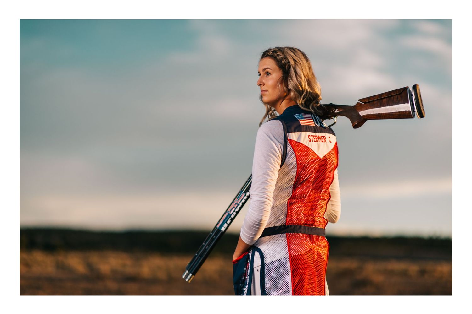 Woman athlete in colorful uniform carries a shotgun over her shoulder outdoors in a field under a cloudy sky.