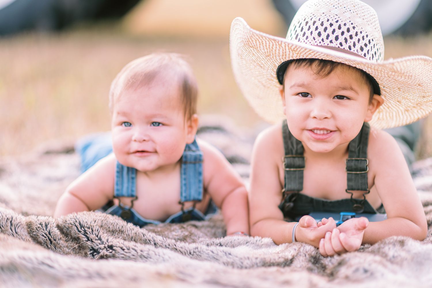 Two cute babies in denim overalls lying on a blanket outdoors, one wearing a straw cowboy hat, smiling at the camera.