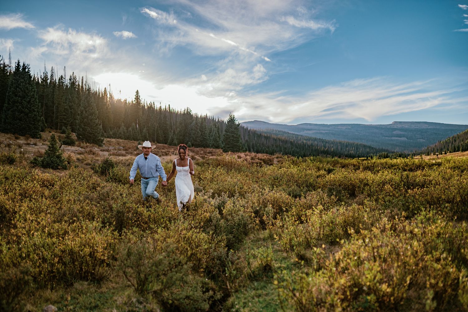 Couple in wedding attire holds hands walking through mountain meadow with evergreen forest and scenic valley at sunset.
