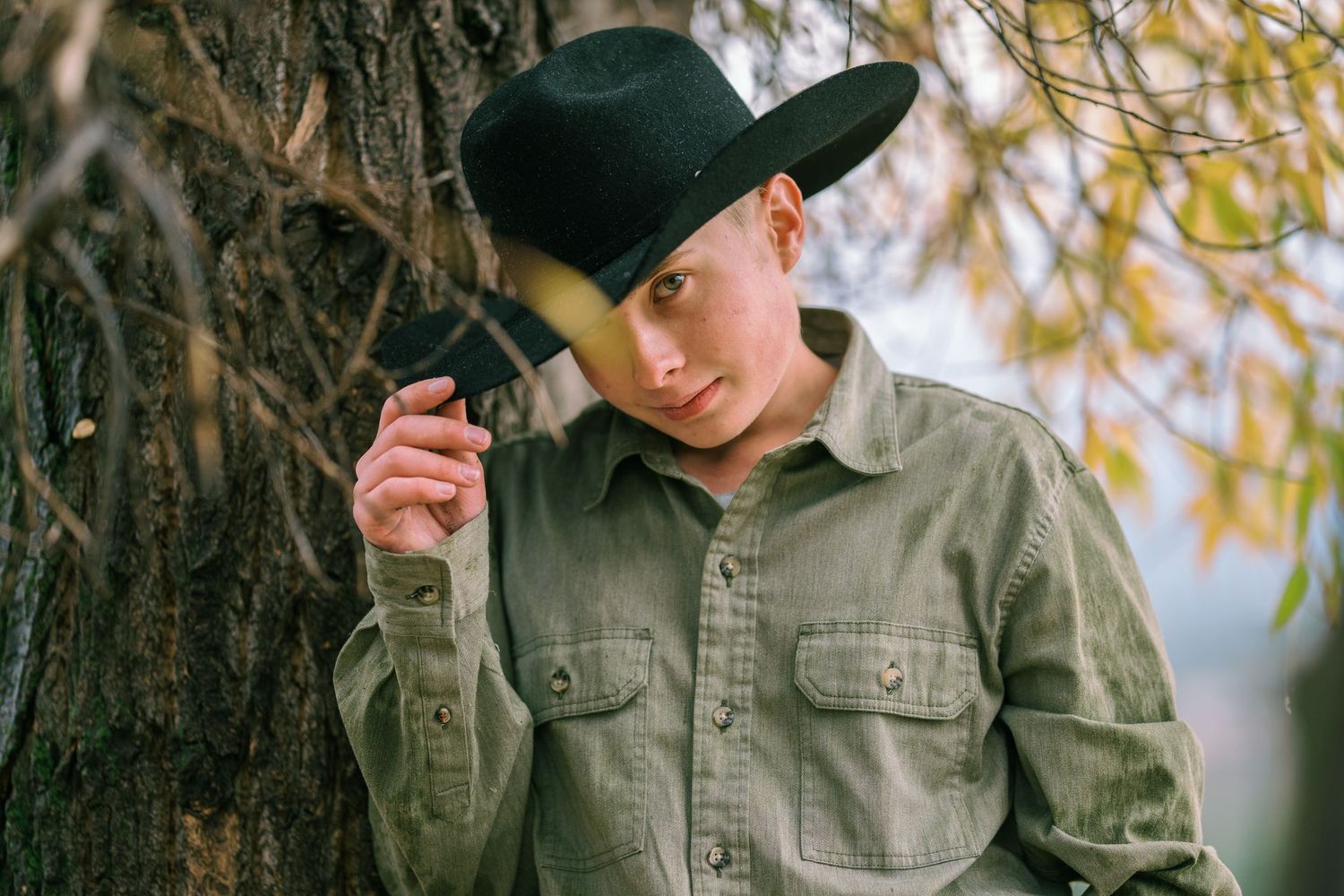 A person in a black cowboy hat and olive green button-up shirt leans against a tree trunk in autumn lighting.