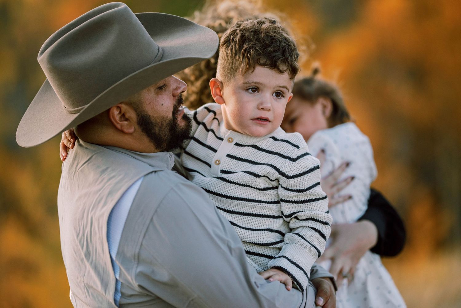 A person in a cowboy hat holding a young child wearing a striped shirt during a warm sunset in nature.