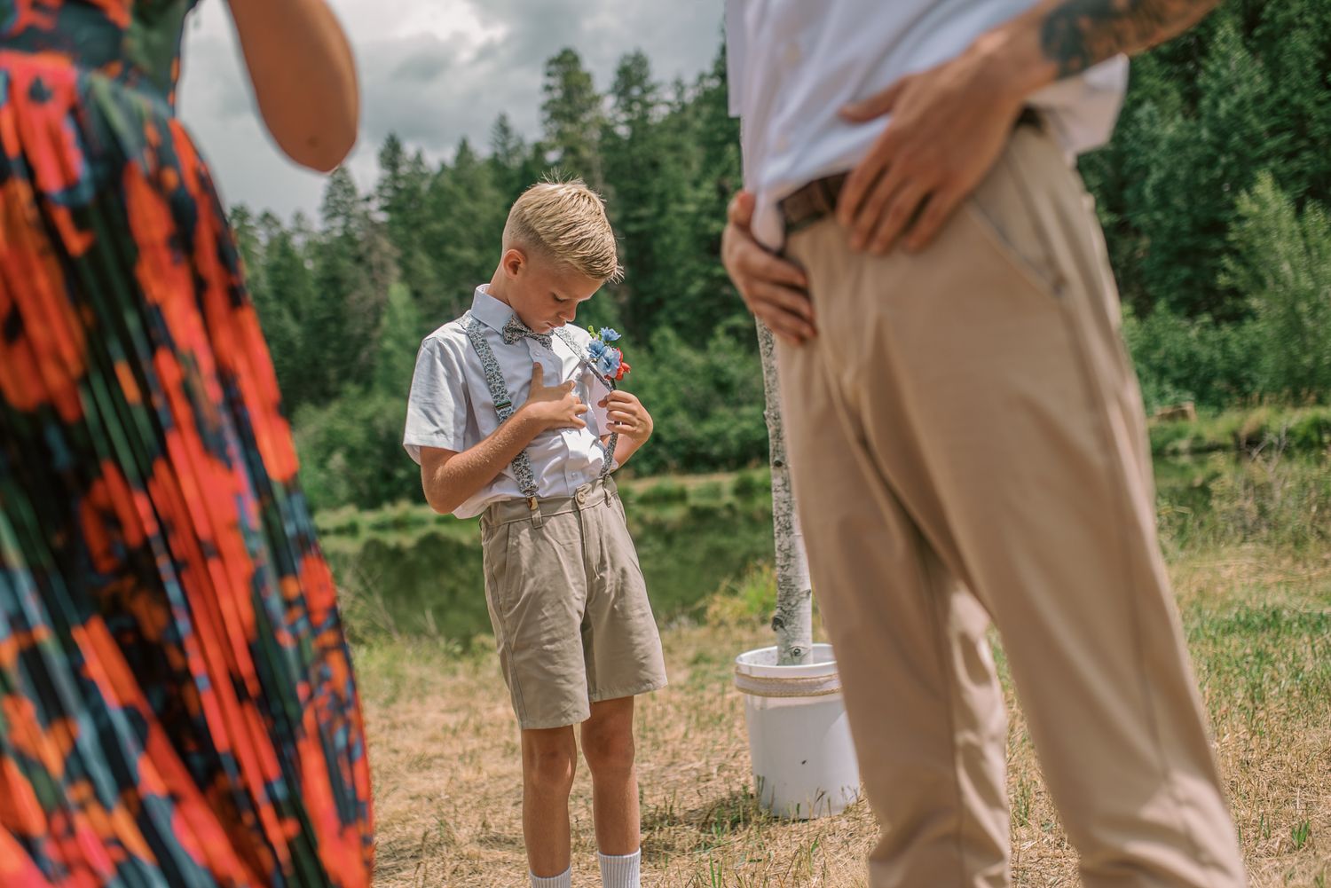 Blonde boy in beige shorts reads paper outdoors near lake, surrounded by adults in colorful attire on sunny day.