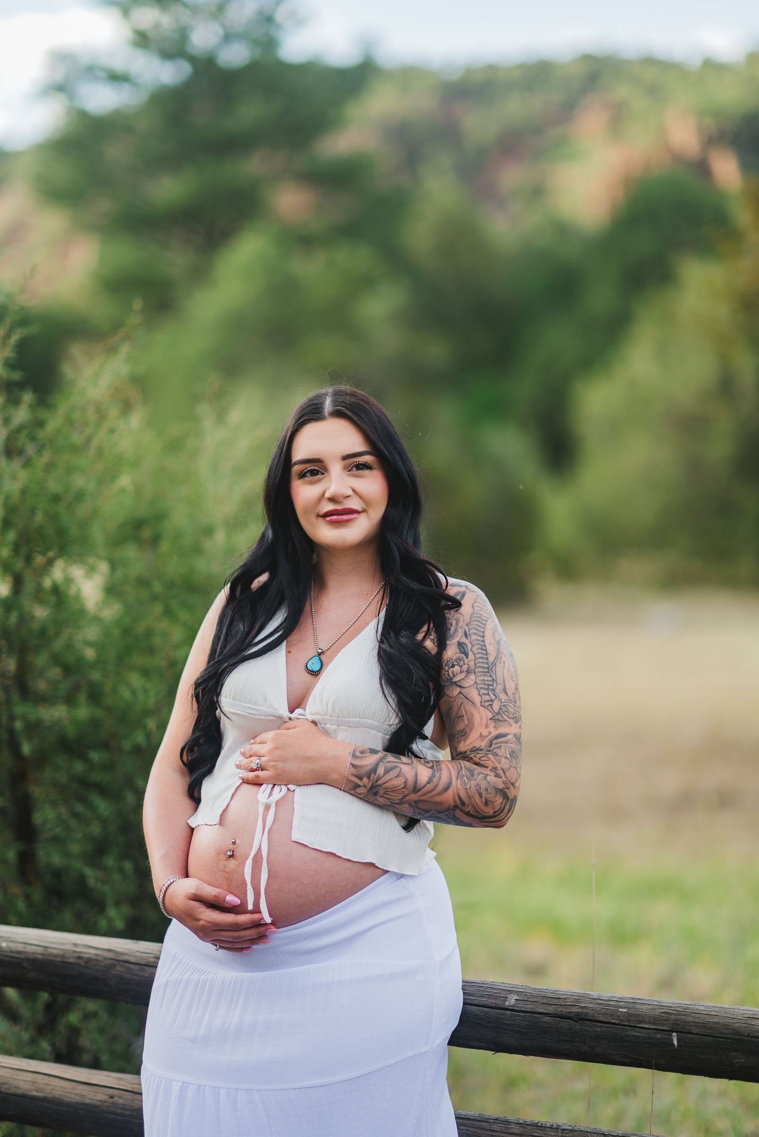 Pregnant woman in white dress with tattoos holds baby bump outdoors near wooden fence with green hills behind her.